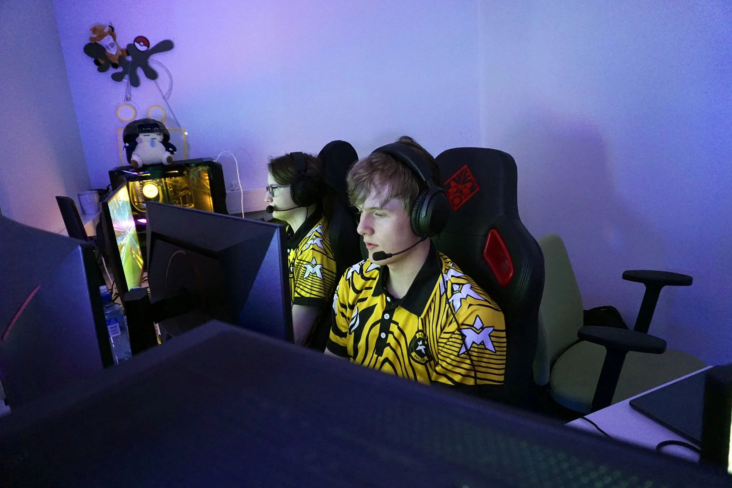 To unge personer som spiller dataspill, begge iført gult og svart kamouflage-liknende drakter, med hodesett på, i et rom med flere datamaskiner.