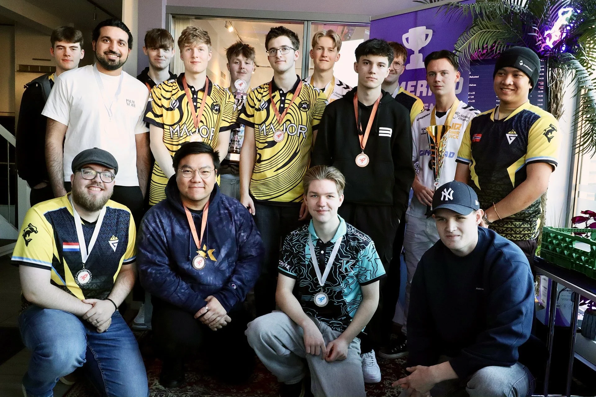 En gruppe unge menn, noen iført gul og svart sportsklær, står og sitter i et rom med grønne planter. De smiler, noen har medaljer rundt halsen og en har en pokal.