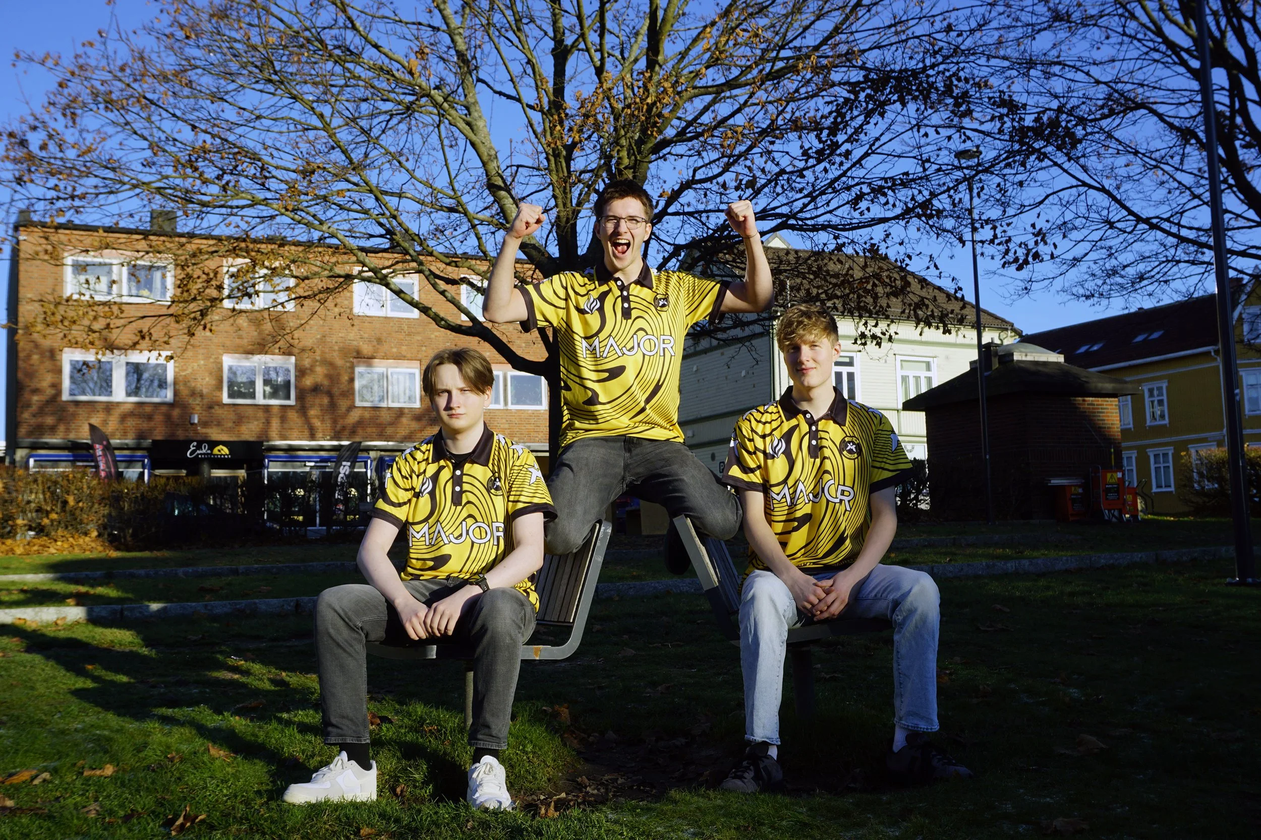 Tre unge menn i gule og svarte fotballdrakter, to av dem sitter på en benk mens den tredjefanser opp med armene i været, på en park med trær og bygninger i bakgrunnen.