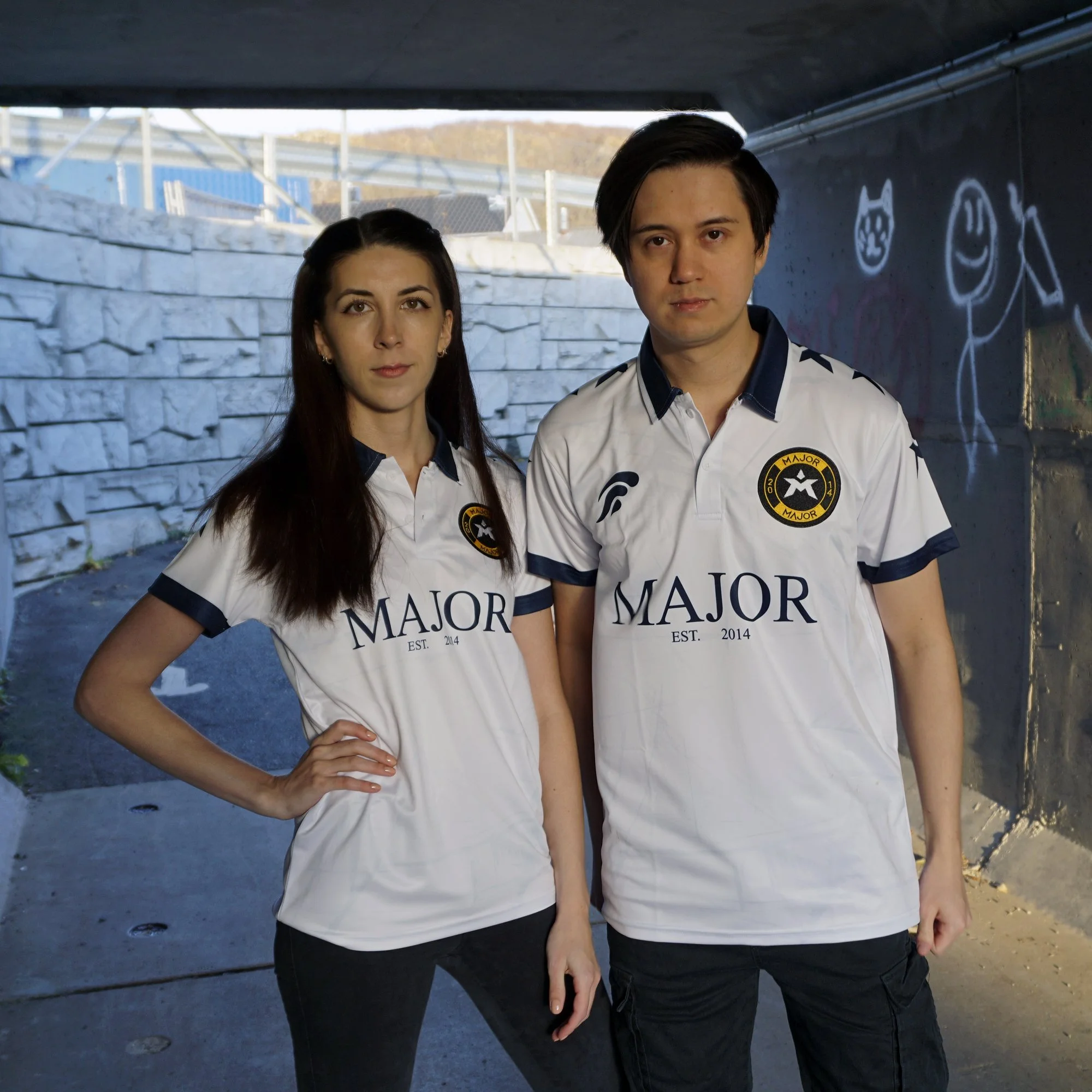 To personer i hvite t-skjorter med logo og teksten 'MAJOR' foran en murvegg med graffiti, under en bro eller tunnel.