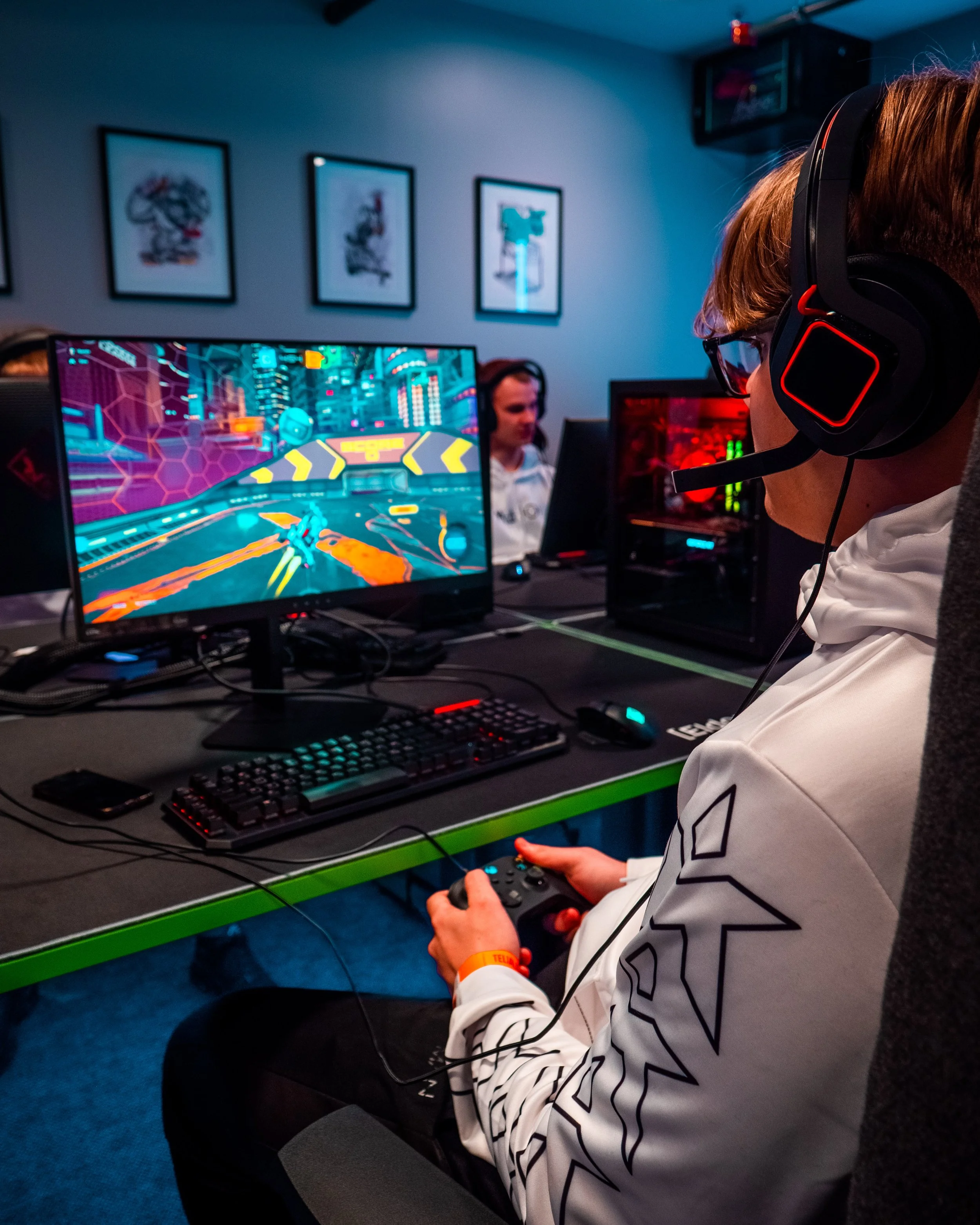 Person som spiller dataspill på en datamaskin i et rom med tre personer som ser på skjermer, omgitt av bilder på veggen.