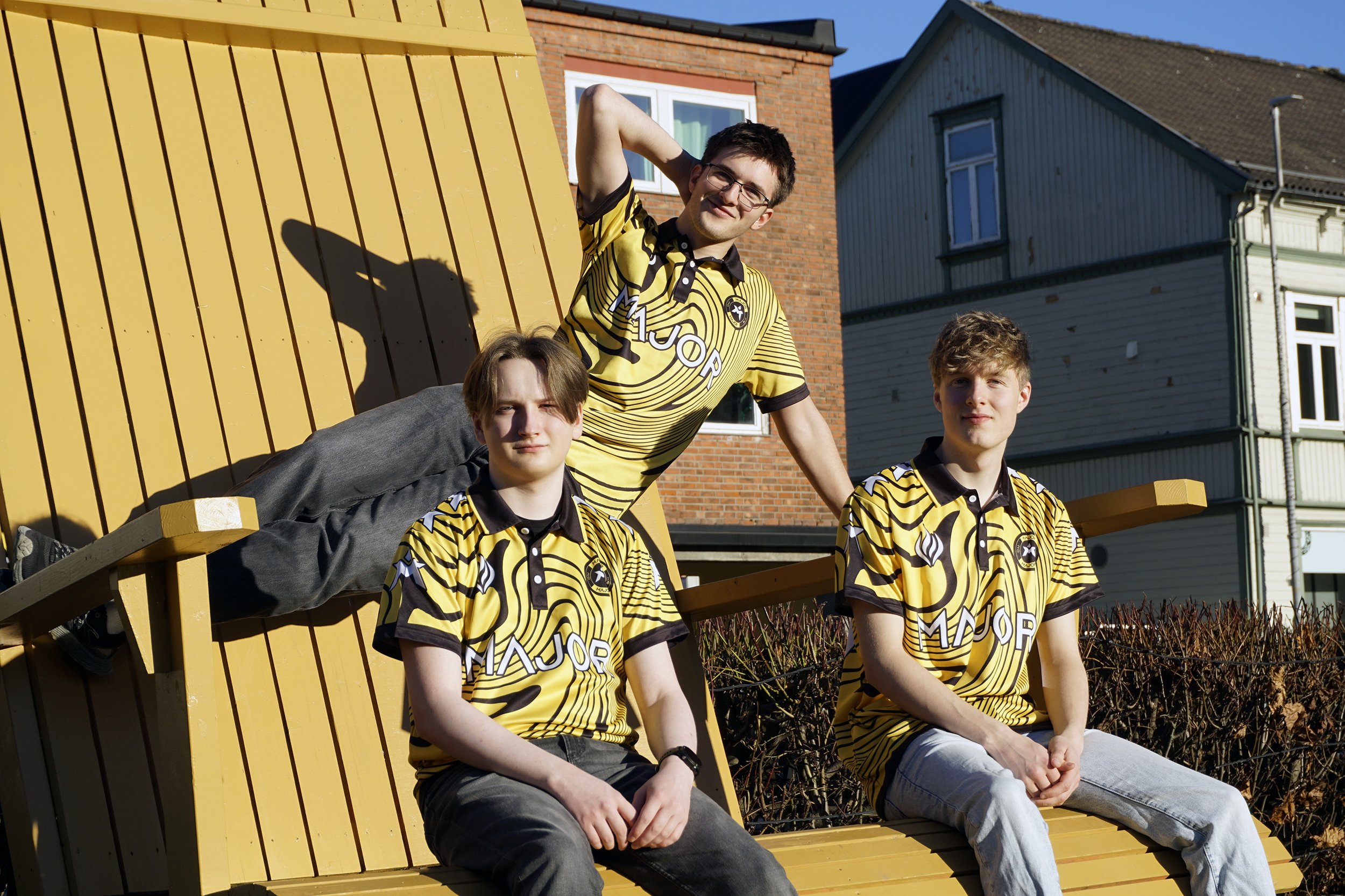 Tre unge menn i gule og svarte-striped sportskjorter som sitter og ligger på en trebenk utendørs på en solrik dag, med hus i bakgrunnen.