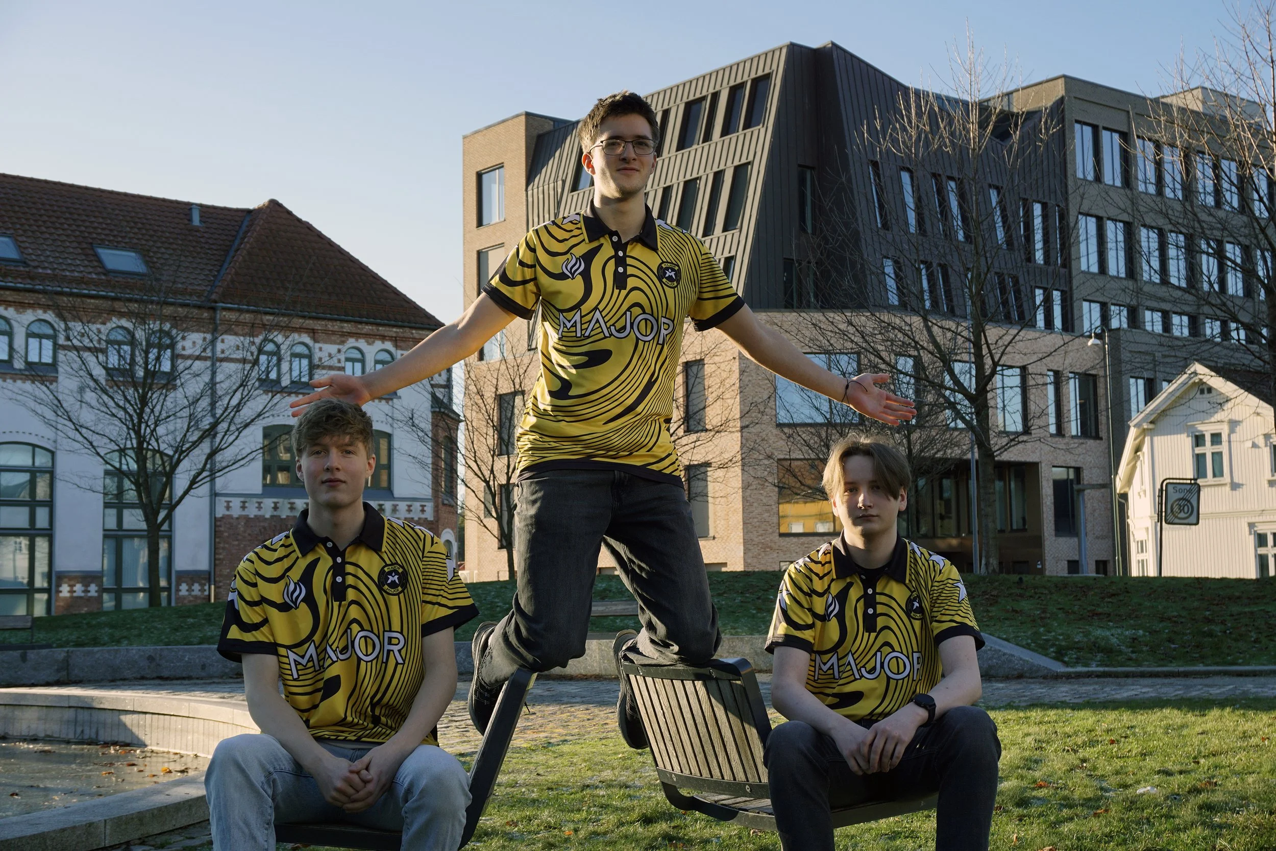 Tre unge menn som sitter og står på en park-benker, alle iført gule og svarte striped t-skjorter med logoen 'Major'.