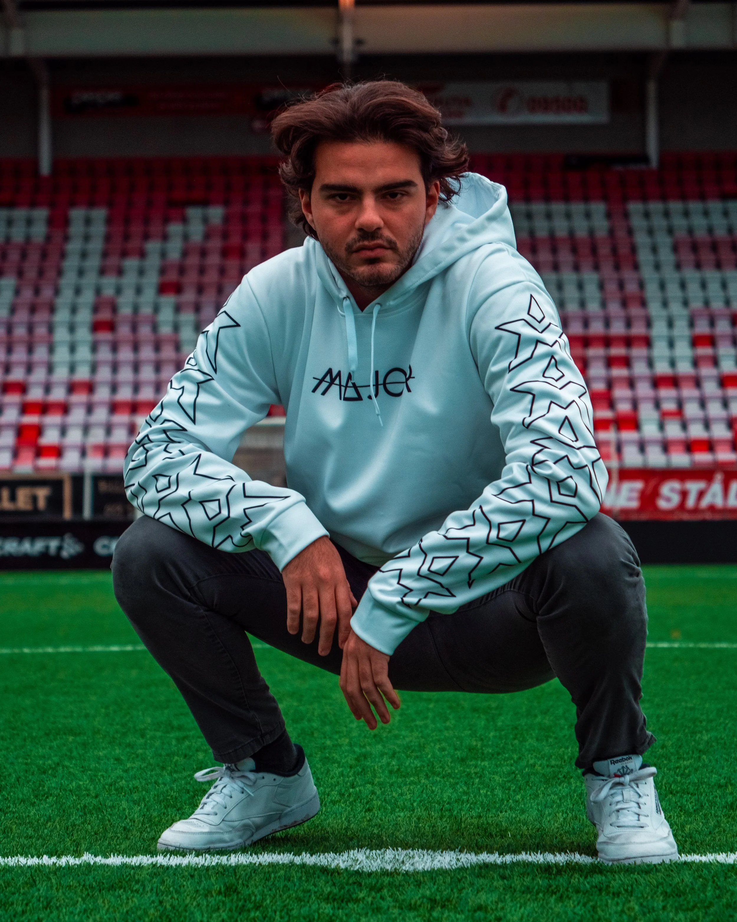 En person i en lys blå hettegenser, brun hår, croucher på en fotballbane.