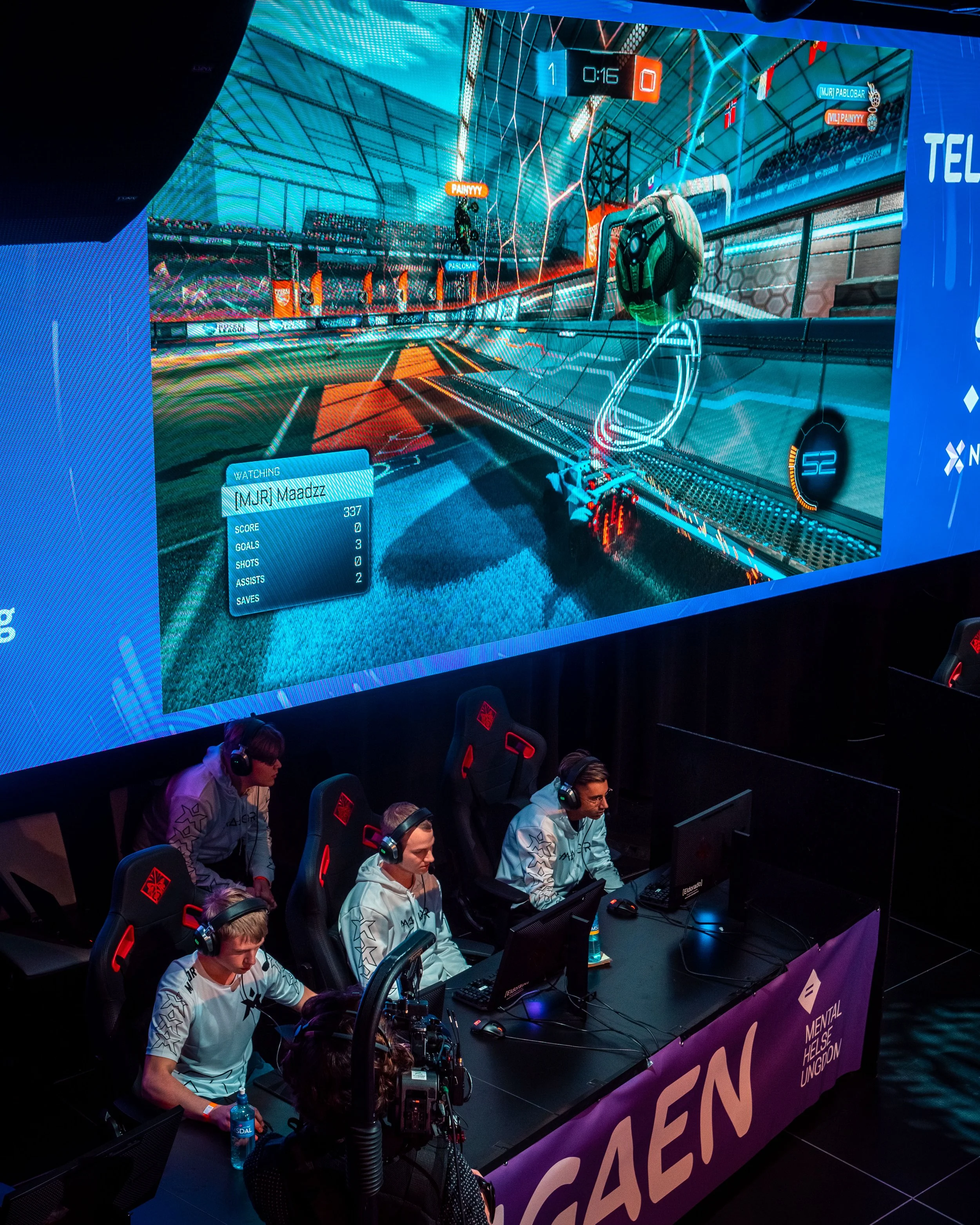 E-sports konkurranse med fire spillere som spiller Rocket League på datamaskiner, mens en stor skjerm bak viser spillets grafikk og score.