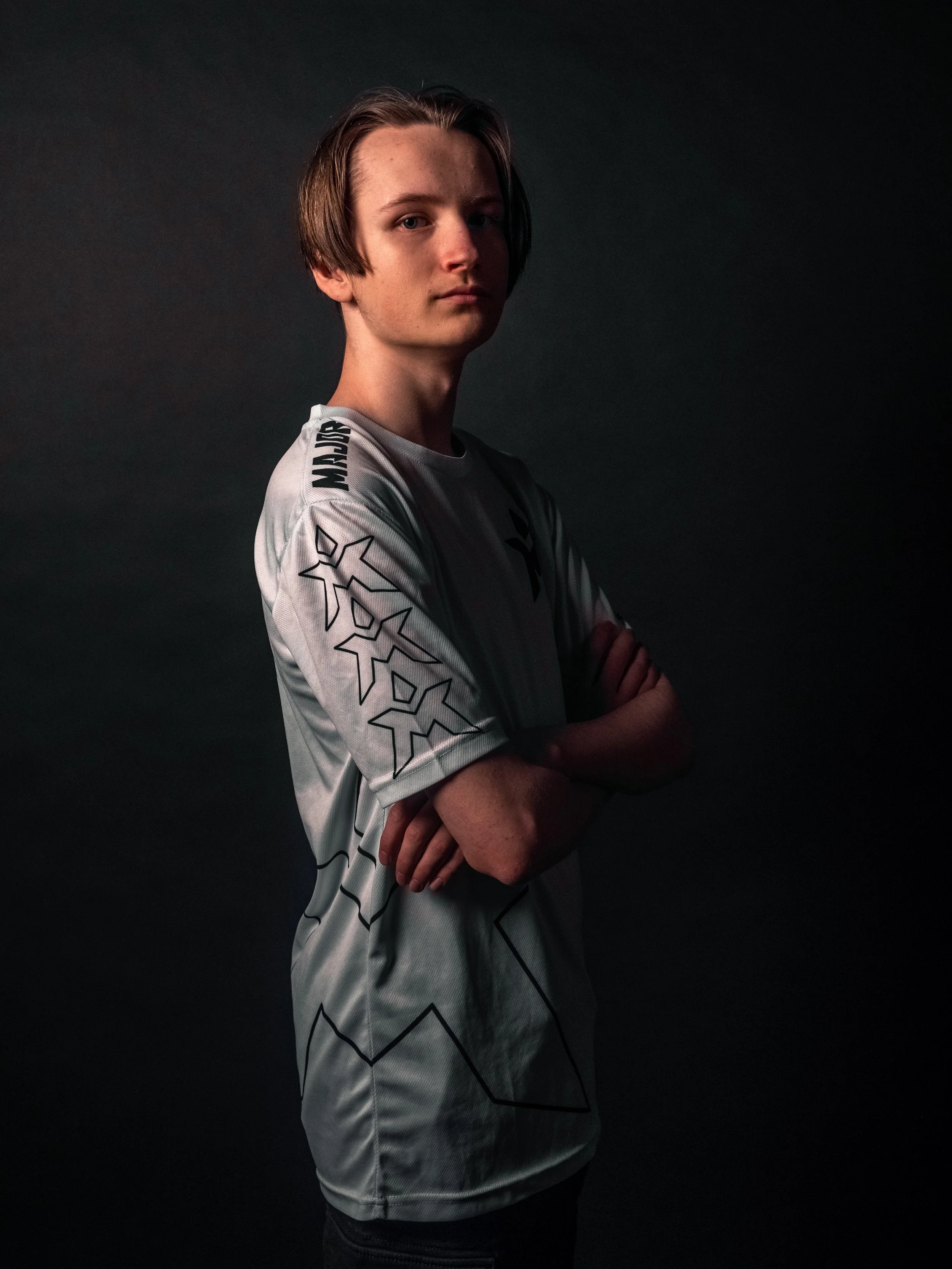 Ung mann med langt, nedoverbøyd hår, kledd i en lys grå sportskjorte med svarte linjer og stjerner på ermer, står med armene krysset foran en mørk bakgrunn.