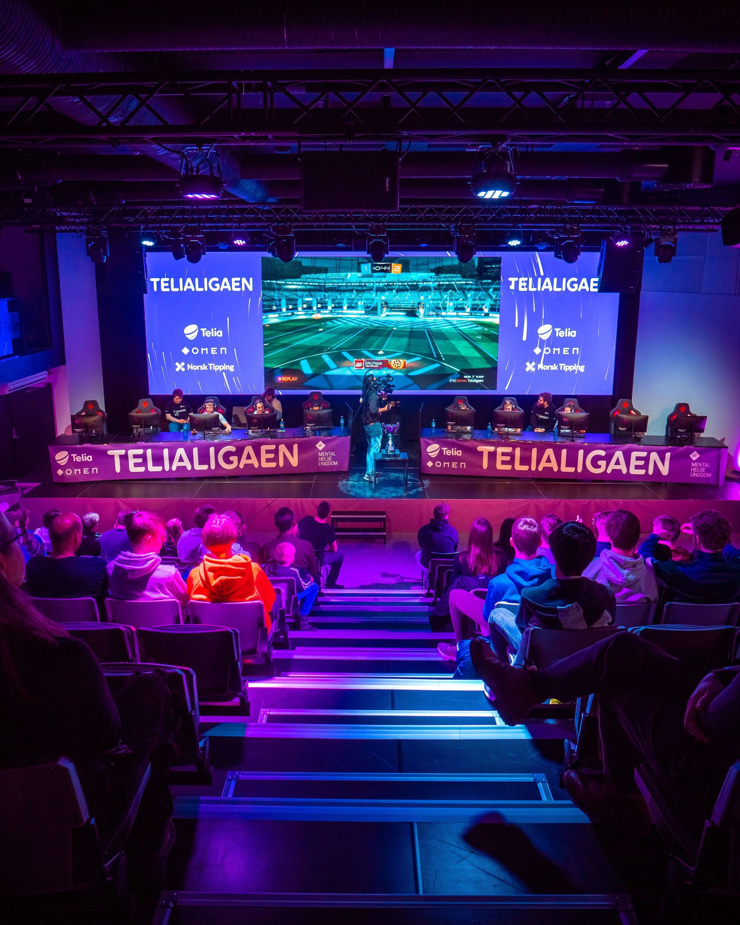 En scene med en gamersamling som spiller e-sport, med store skjermer som viser et videospill, og et publikum som ser på.