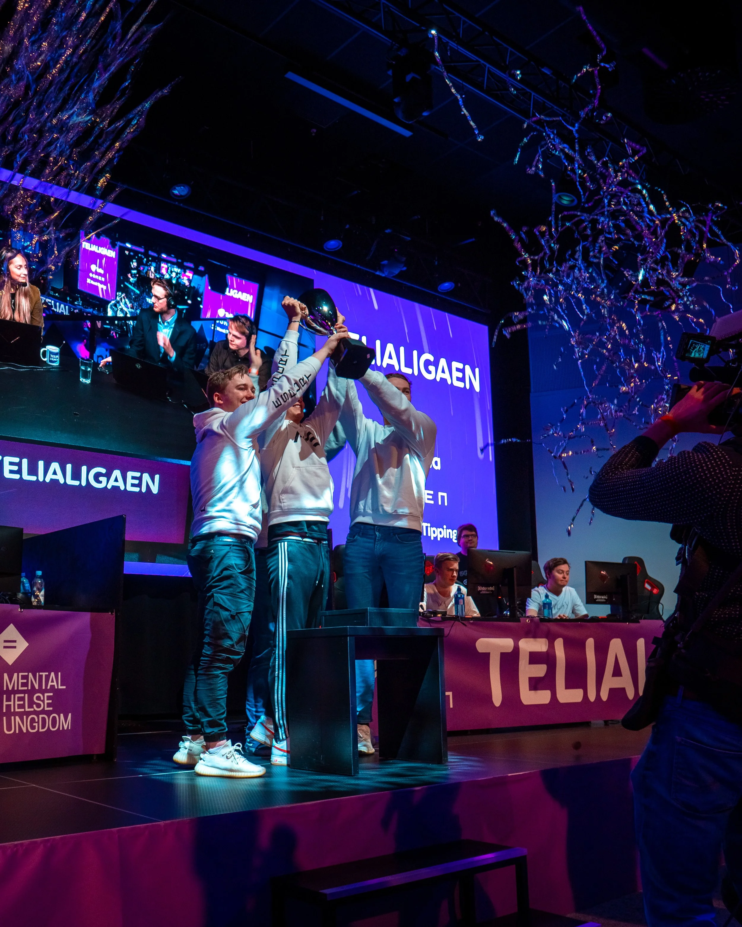 Tre personer står på en scene og holder opp en pokal sammen, mens andre ser på. Bak dem er en stor skjerm med teksten 'TELIALIGAEN'. Det er dekorasjoner og konfetti i luften, og personer foran scenen filmer eller tar bilder.