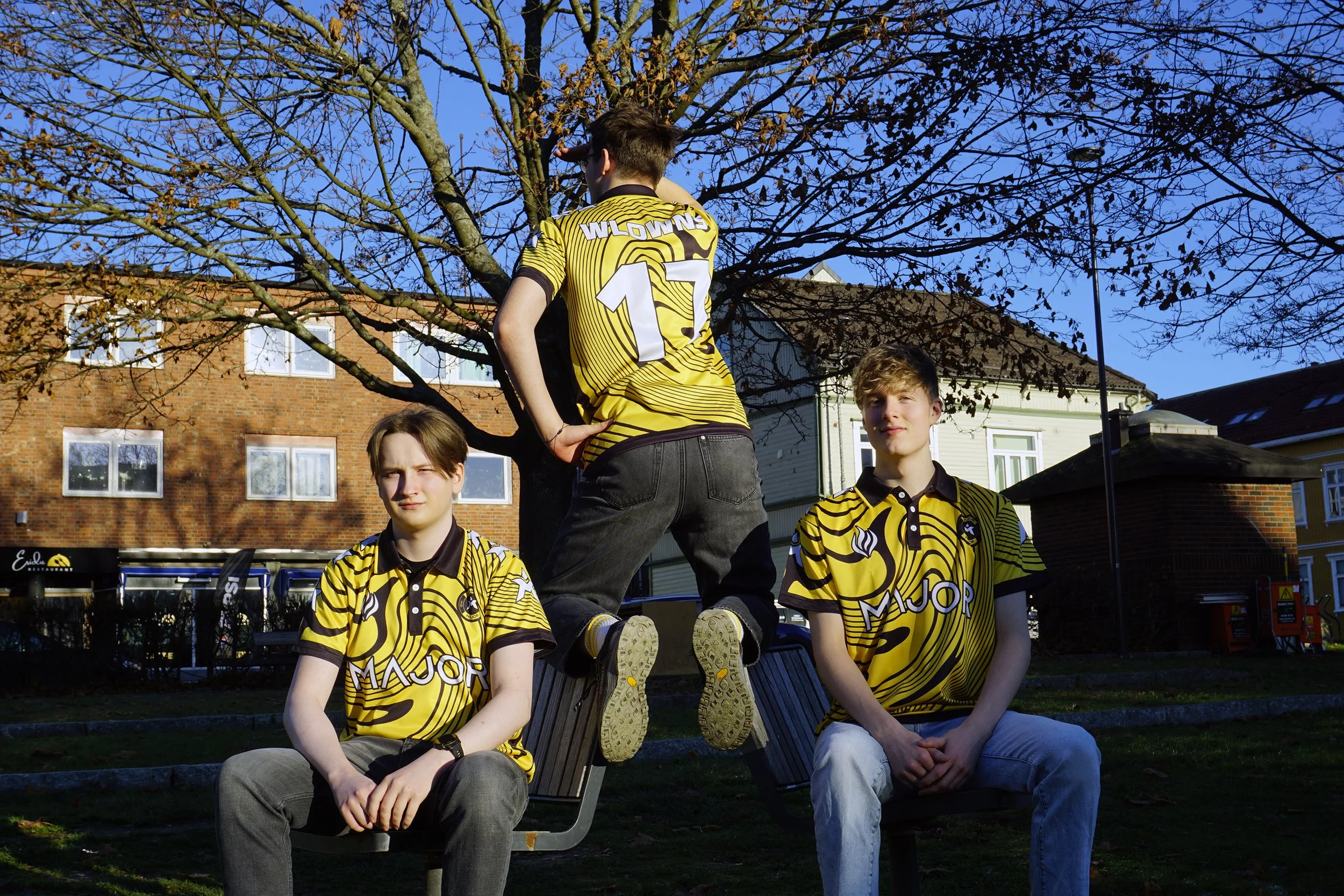 Tre ungdommer i gule og svarte striped fotballdrakter med logo og tekst, to sitter på benker og en står på bukker, foran en trærerr med høstblader og bak en bolig med røde murstein og hvite vinduer.