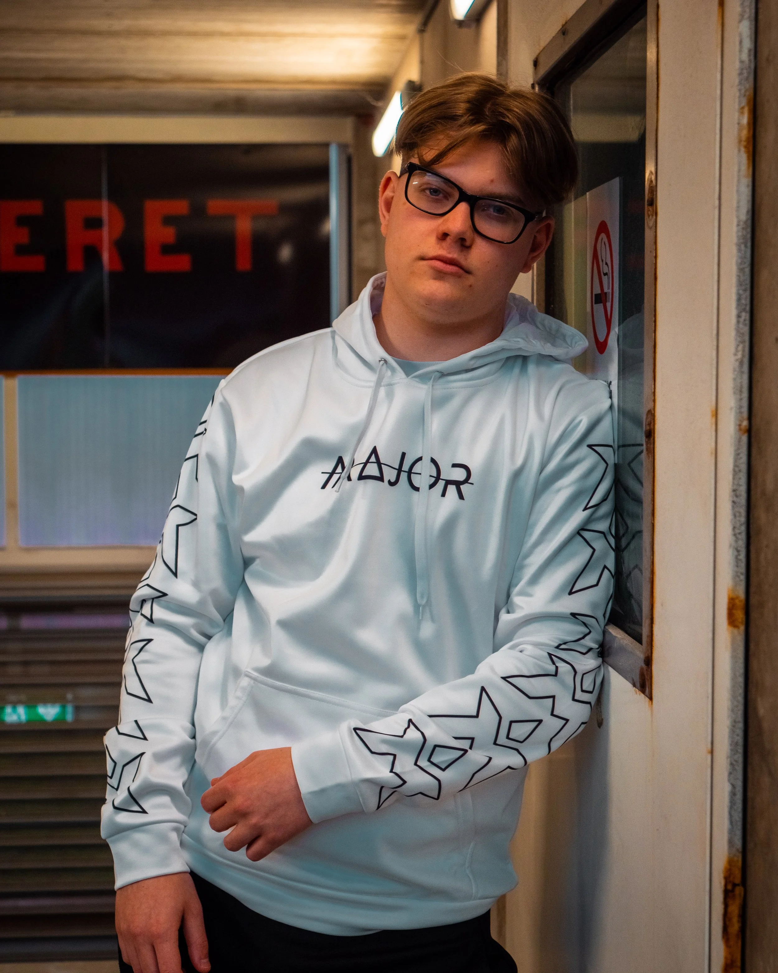 Ung mann med briller, iført en hvit hoodie med svarte geometriske mønstre, står med krysset armer og ser mot kamera i en undergrunnsstasjon.