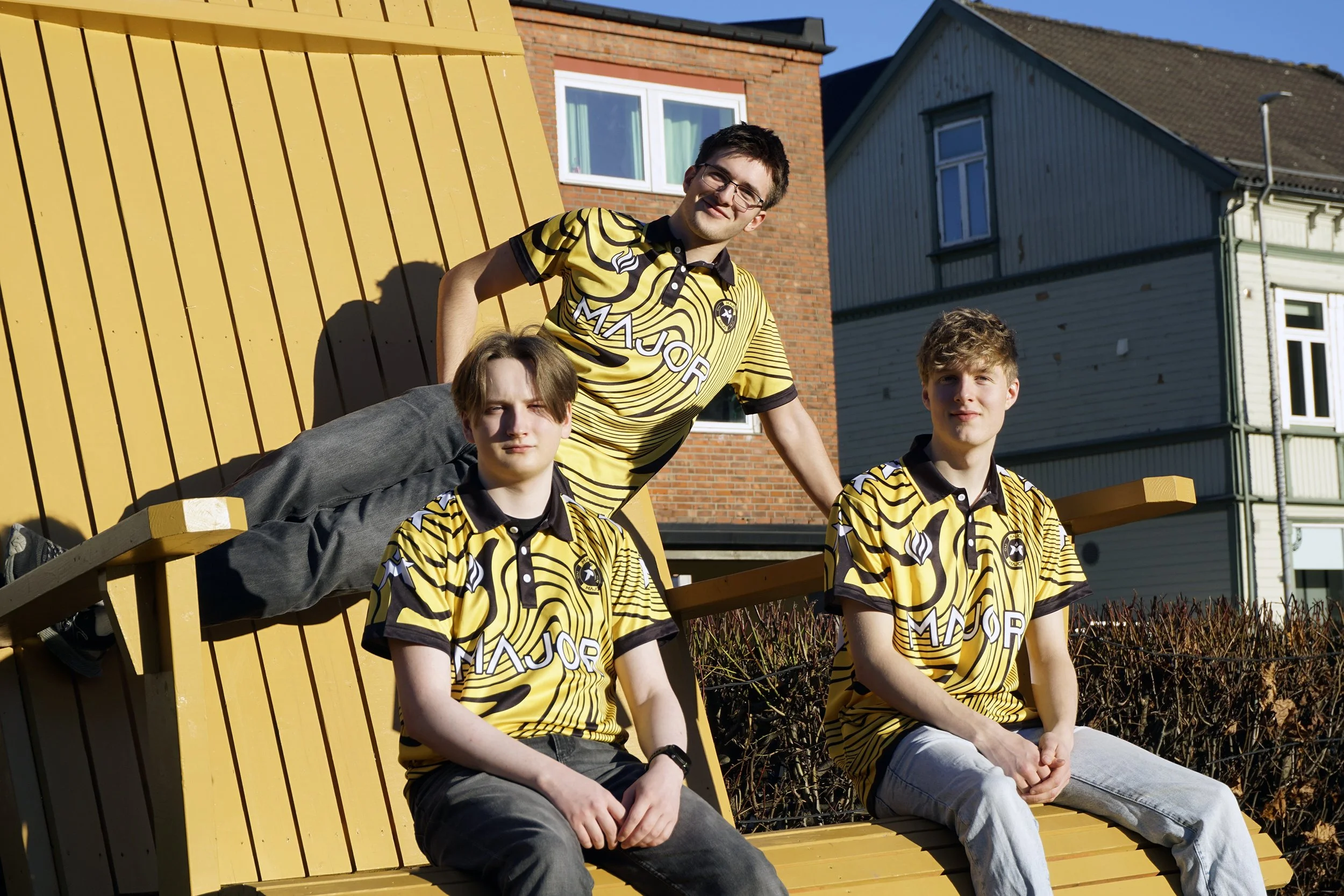 Tre unge menn i gult og svart stripete t-skjorter sitter og ligger på en gul trebenk utenfor. Én ligger på ryggen med hodet på ryggstøtten, mens de to andre sitter foran den. De er på en solrik dag med bakgrunn av hus og busker.