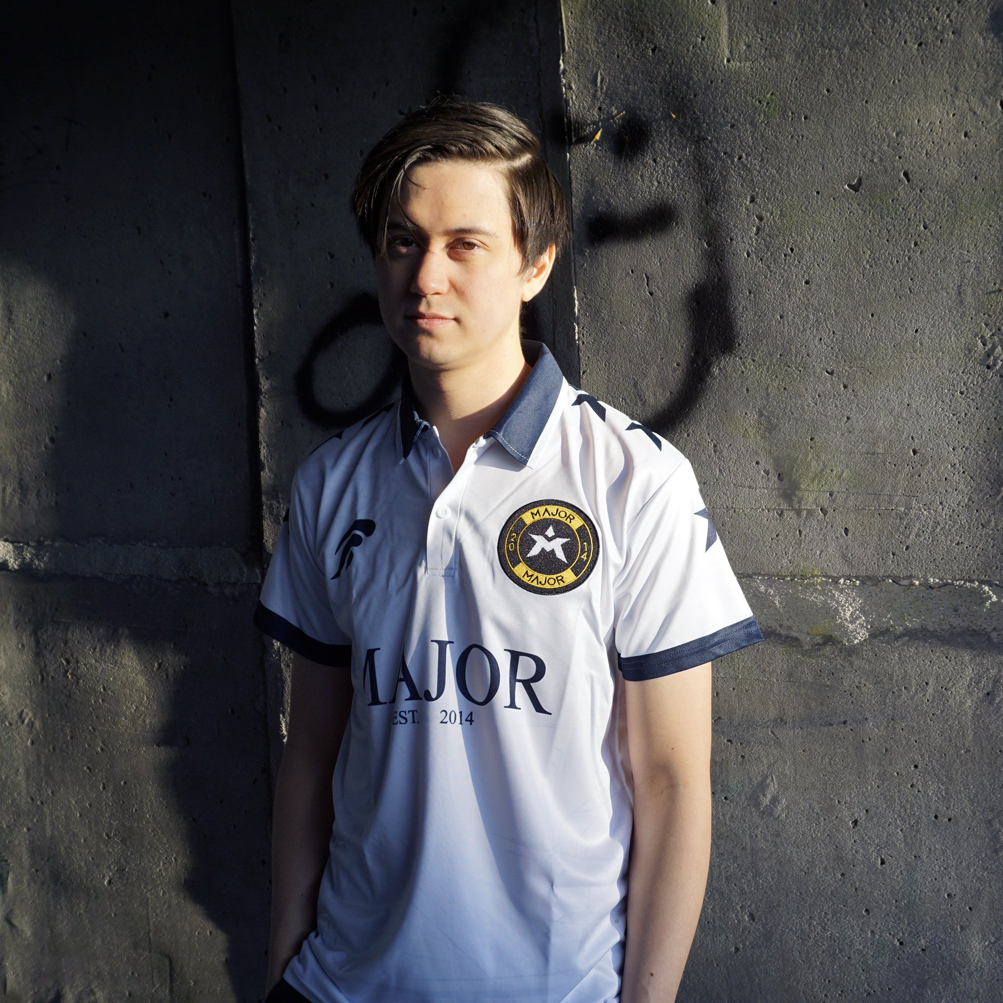 En ung mann med kort, brunt hår står mot en vegg, iført en hvit og blå sportskjorte med en rund logo på brystet og teksten 'Major'.