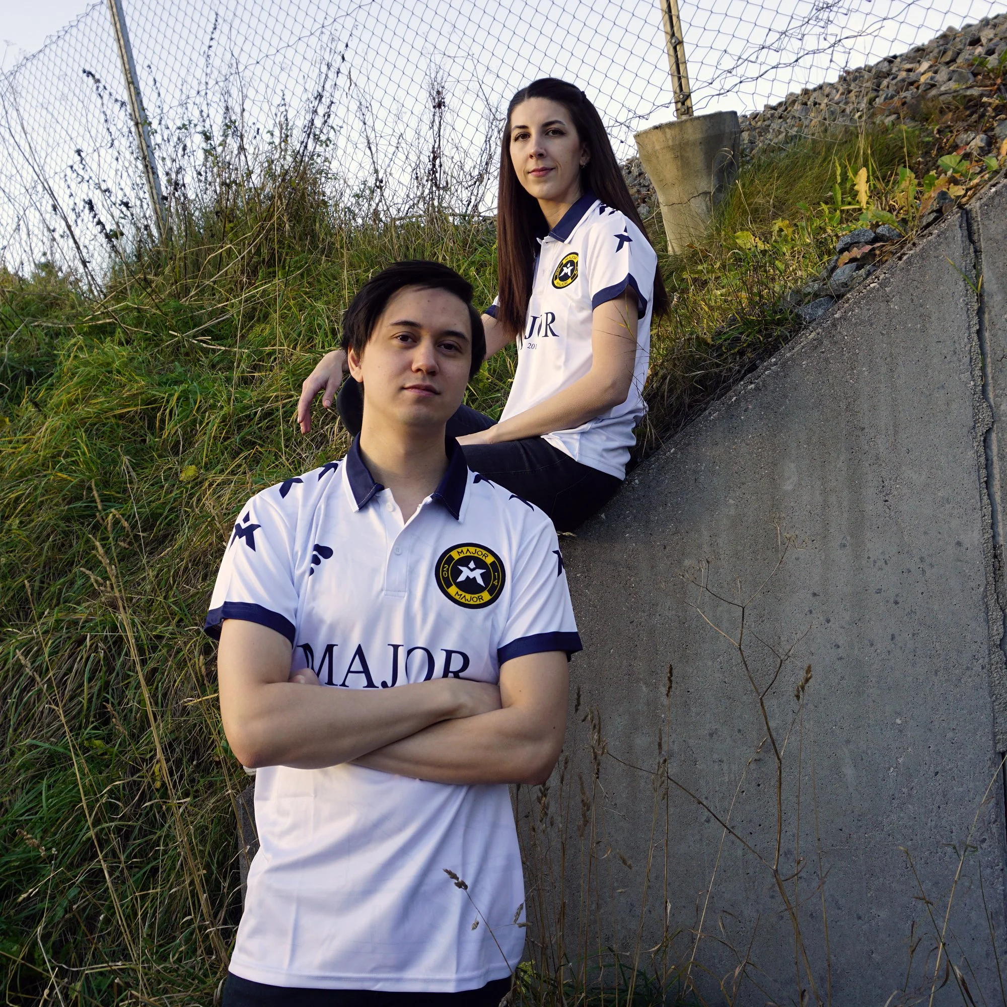 To personer iført hvite fotballdrakter med logo og tekst, foran en grønnskridd bakke og en betongmur.