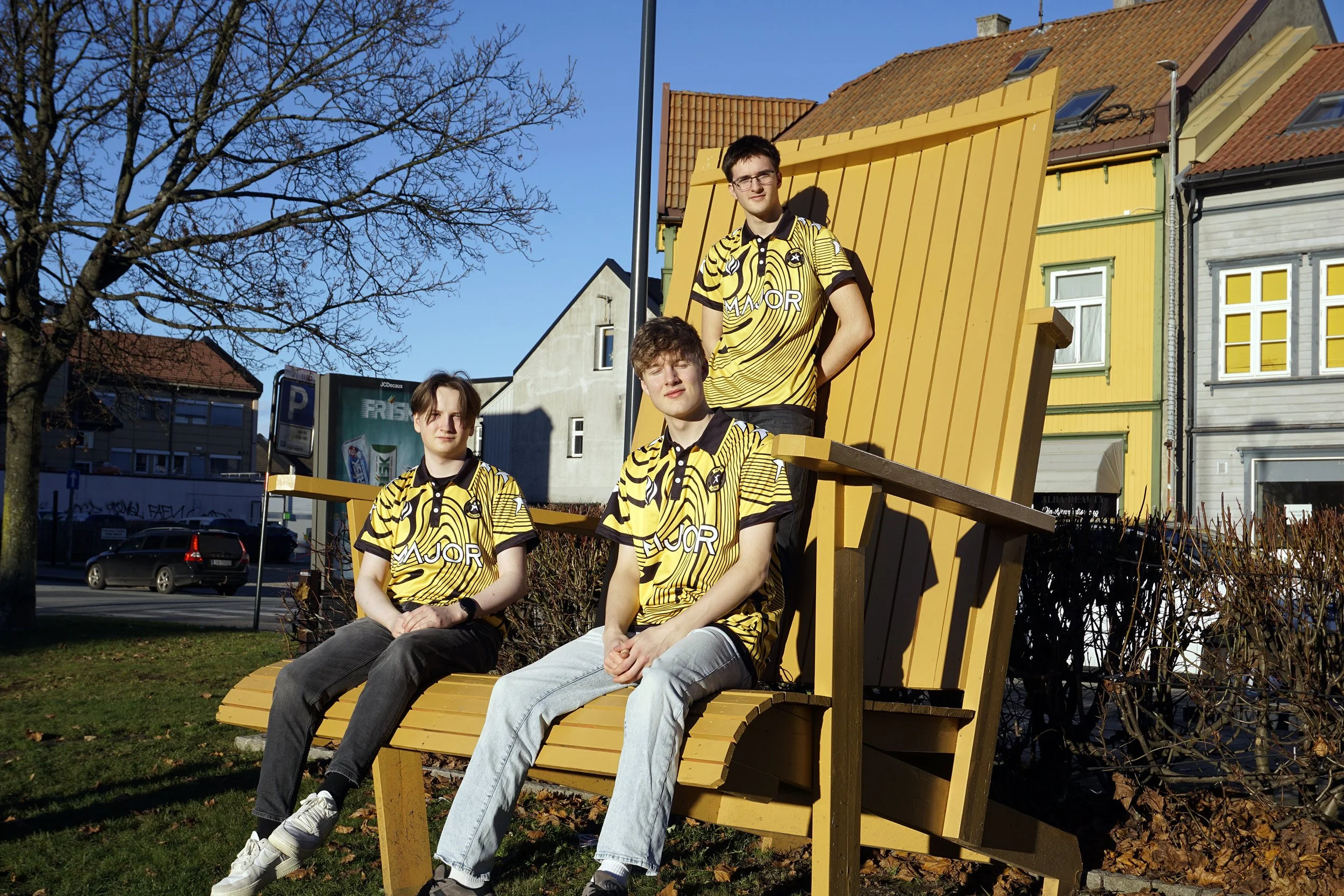 Tre personer som sitter og står på en stor trestolsseng utendørs, alle iført gule og svarte sportsklær, mot en bakgrunn av fargerike hus og en blå himmel.