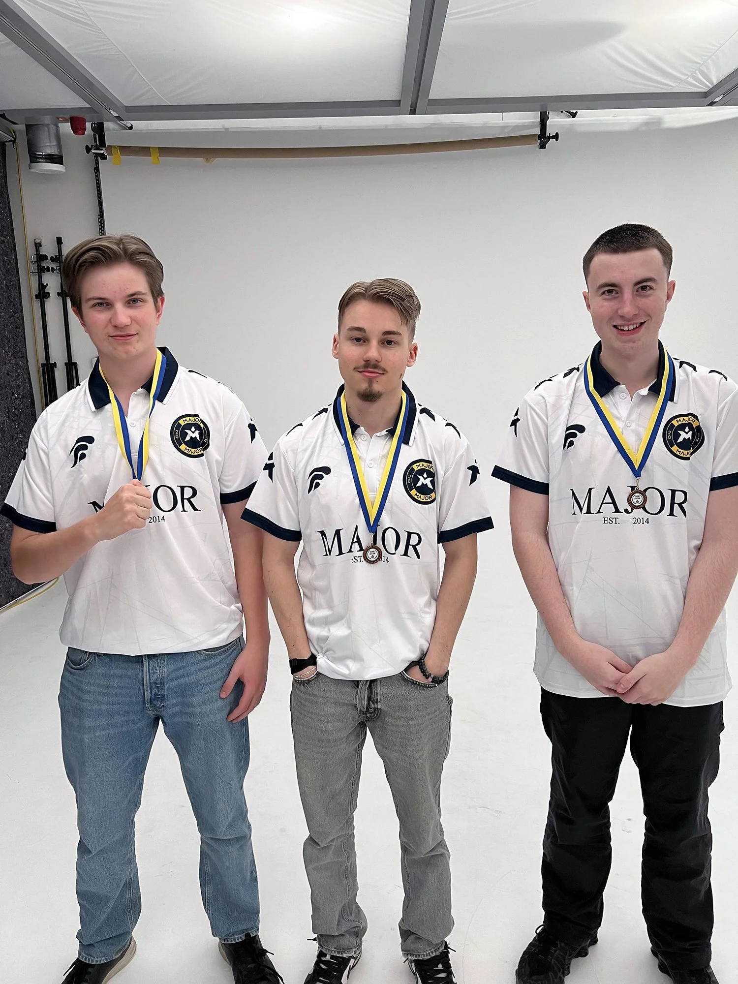 Tre unge menn i hvite sportskjorter med medaljer rundt halsen, står på en fotobakgrunn med en hvit vegg. De smiler og poserer for bilde.