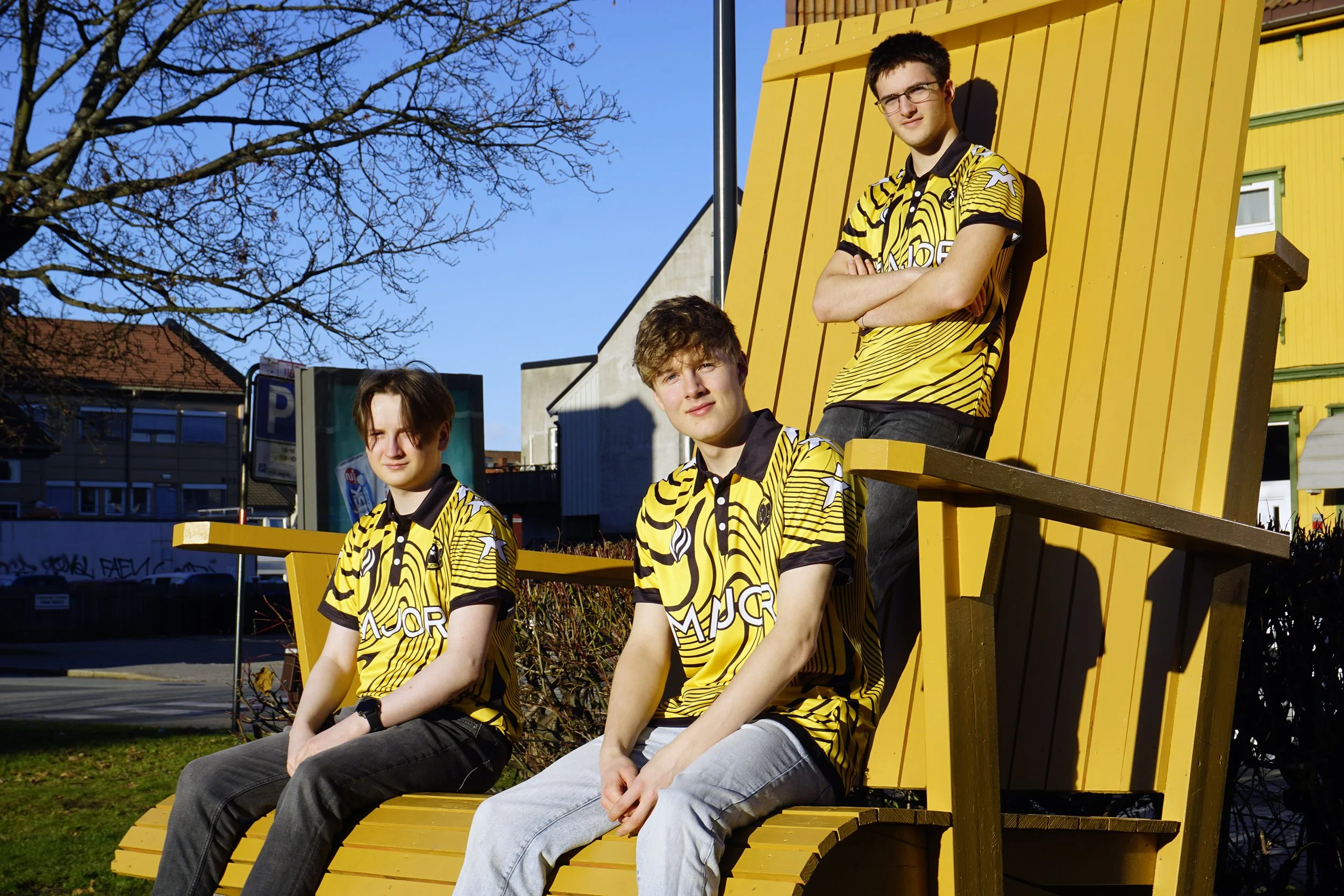 Tre unge menn i gule og svarte fotballdrakter sitter på en stor gul trebenk ute i solen