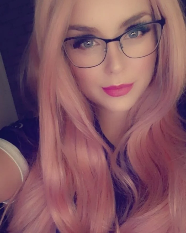 Nærbilde av en kvinne med rosa langt hår, briller, og rødt leppefarge.