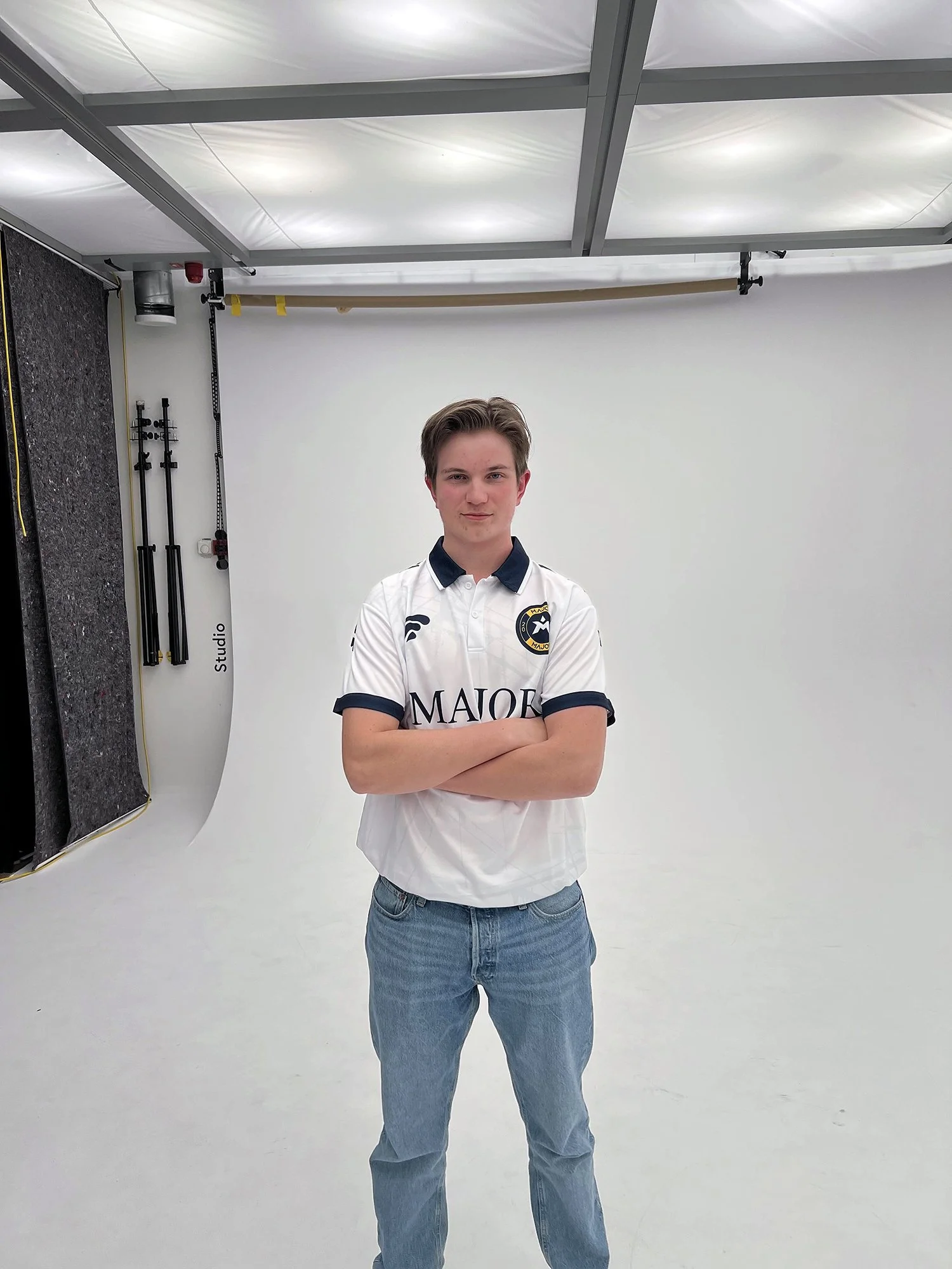 Ung mann står med armene krysset i en fotostudie, iført en hvit fotballdrakt med logo og tekst. Bakgrunnen er en hvit backdrop, og det er studioutstyr synlig til venstre.