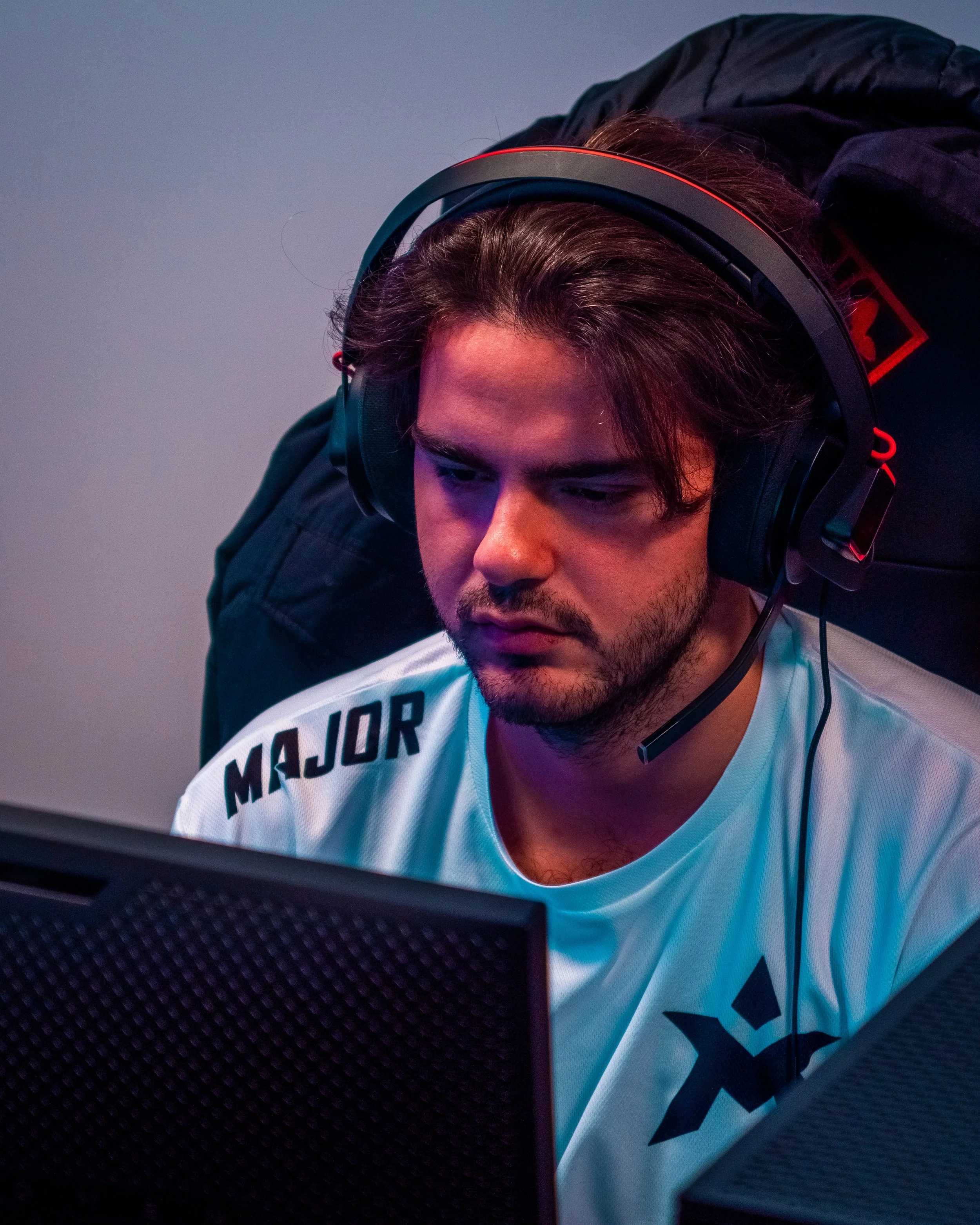 En mann med langt, mørkt hår og skjegg som ser på en dataskjerm, iført gaming-headset og en hvit t-skjorte med ordet "Major".