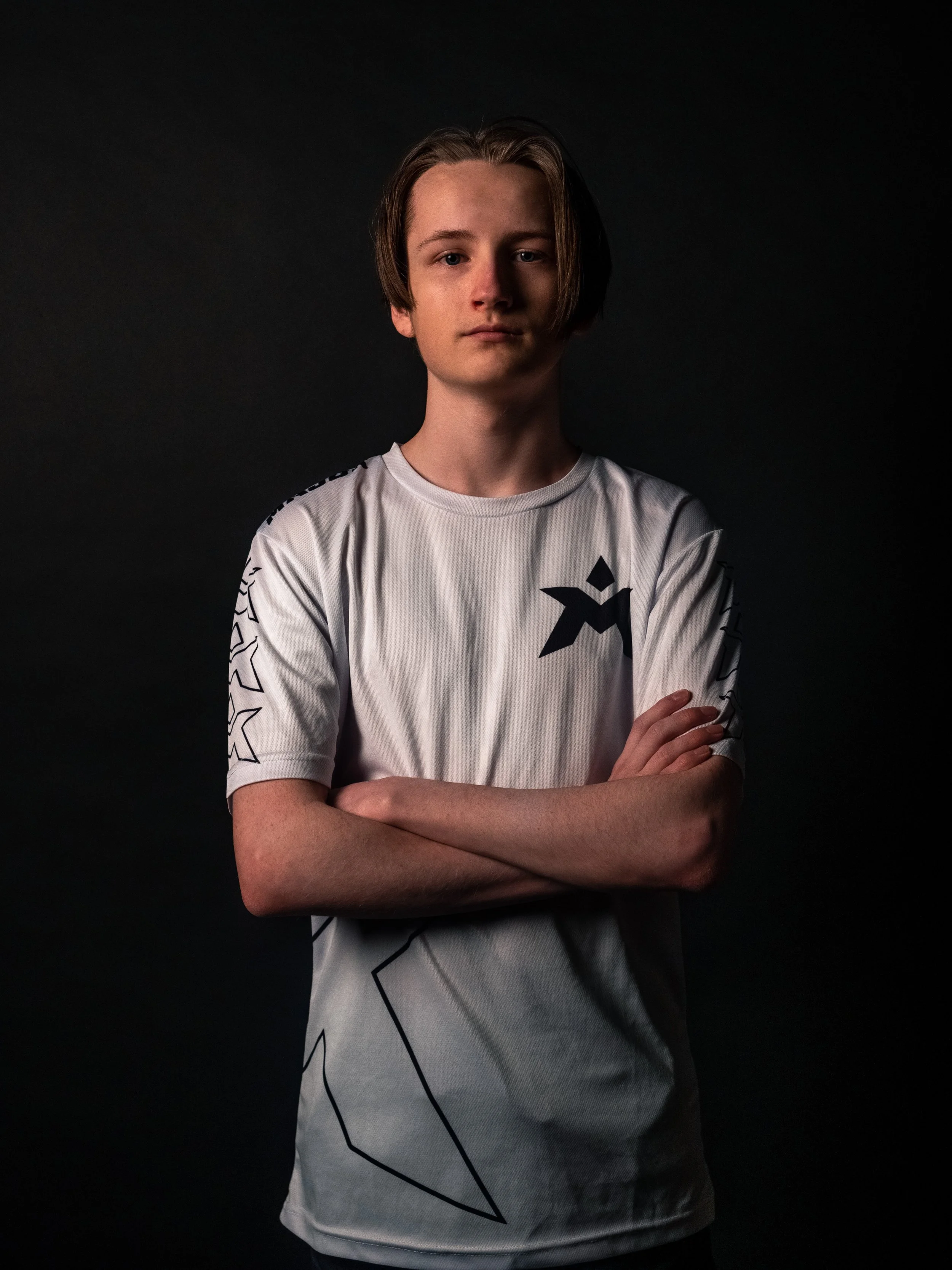 Ung gutt med korslagte armer, kort hår, iført en hvit t-skjorte med svart design.