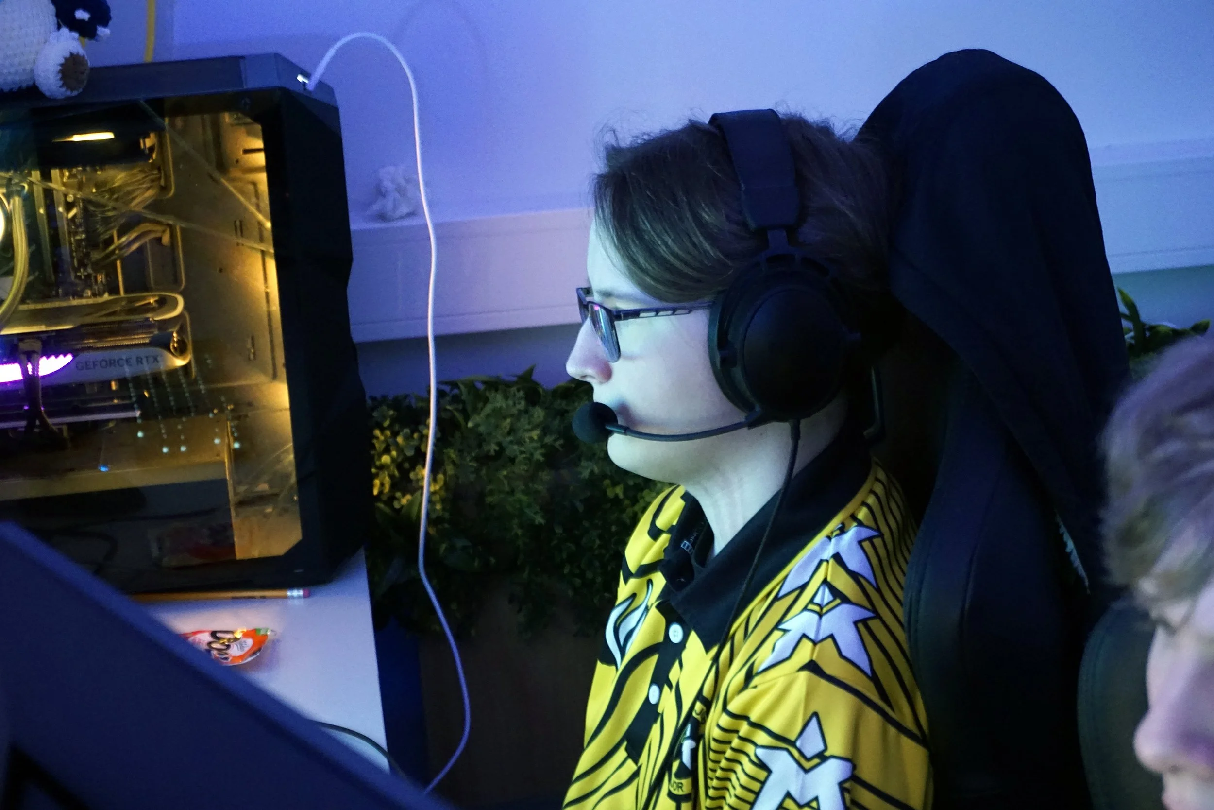 En person med briller og hodetelefoner som jobber ved en datamaskin med synlig grafikkort, med en plante i bakgrunnen.