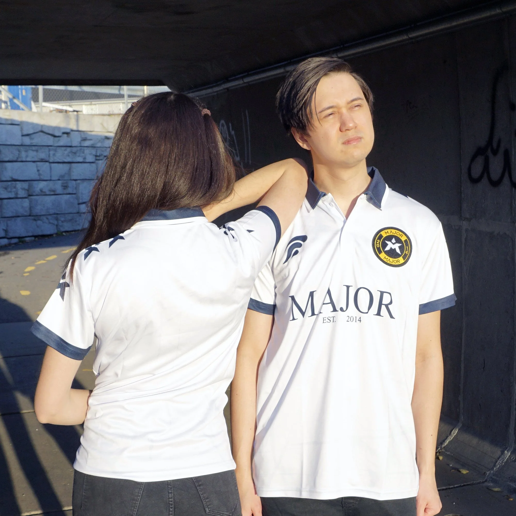 En kvinne i en hvit T-skjorte holder en arm rundt en mann iført en matchende hvit sports T-skjorte, under en bro eller tunnel.