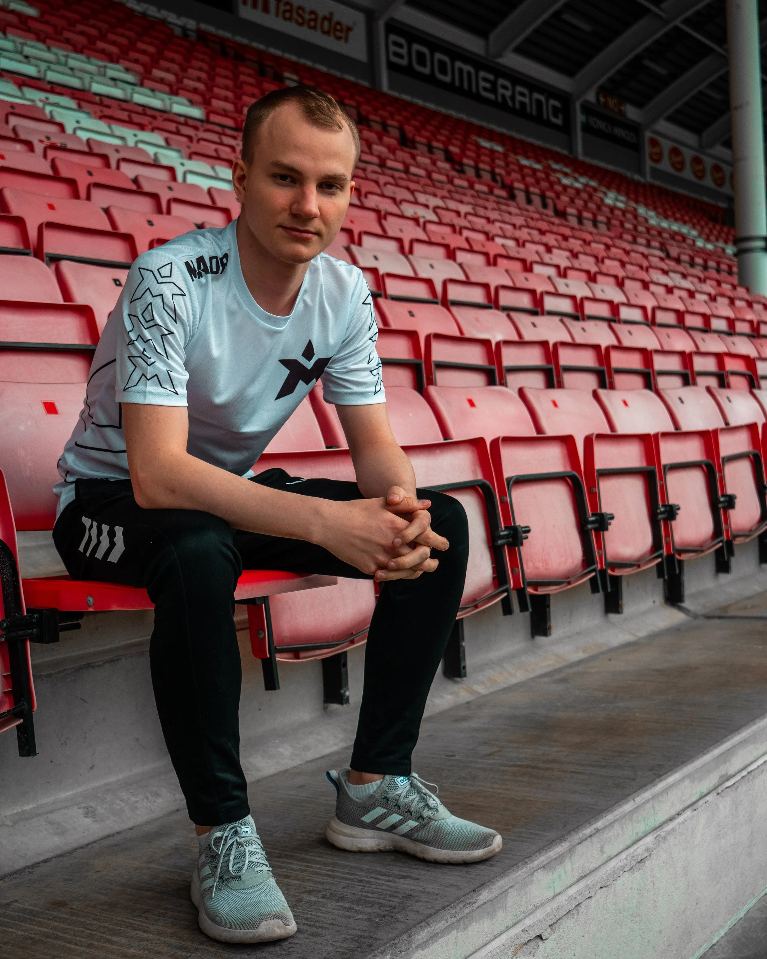 En ung mann sitter på en tom tribuneseats i en sportsarena, iført hvit t-skjorte, svarte joggebukser og grå sportsko.
