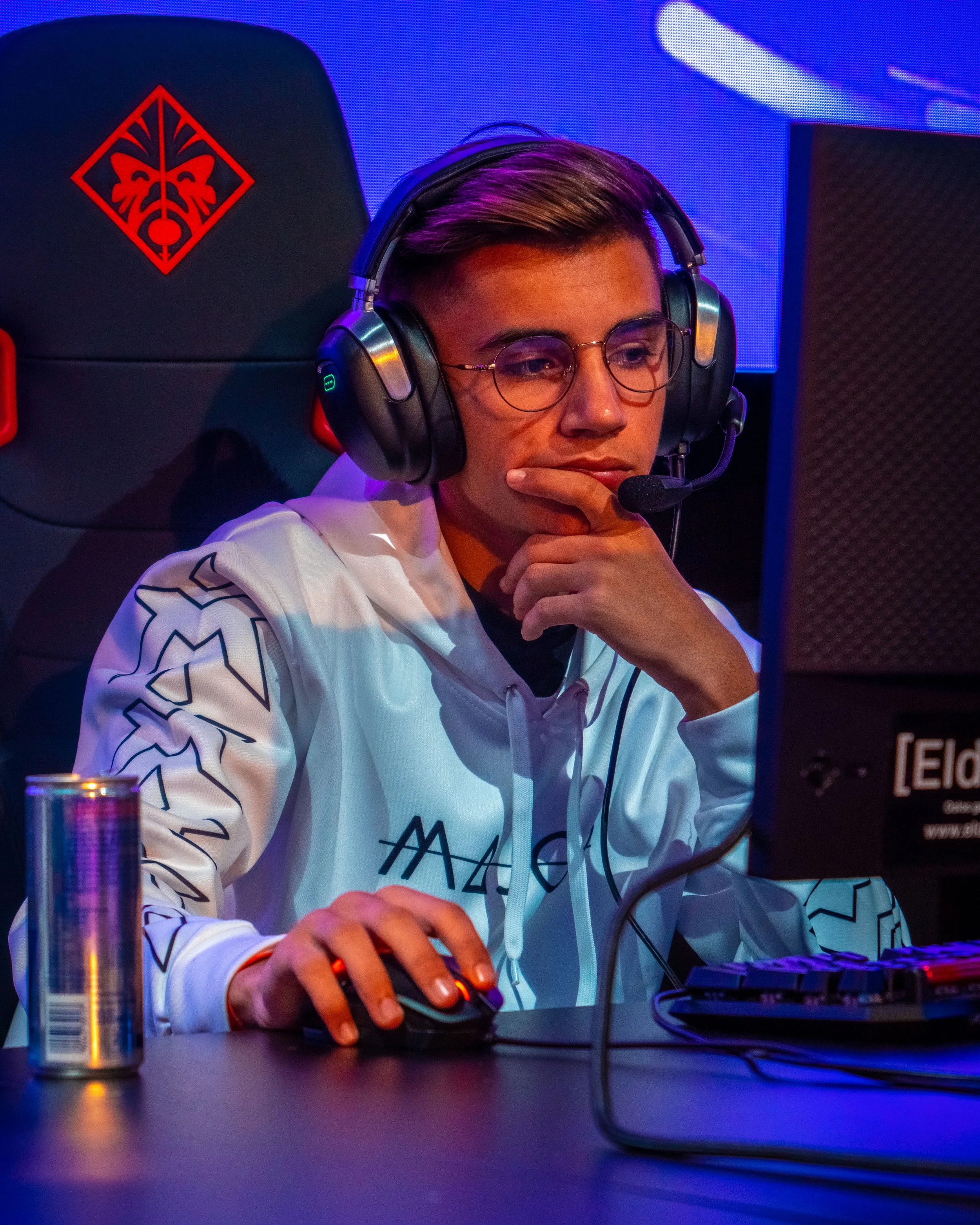 En ung mann med briller og hodetelefoner spiller dataspill, sitter foran en skjerm, med en tankerfull uttrykk, og et energidrikksdåse på bordet.