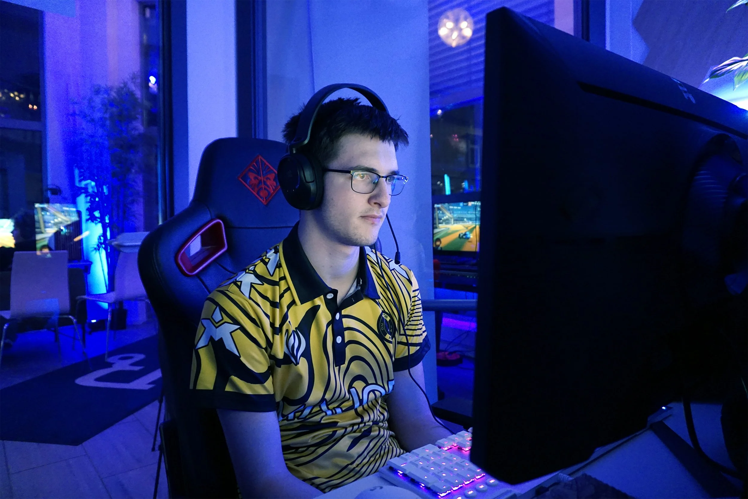 En ung mann med briller og gamingantrekk spiller dataspill ved et skrivebord i et gamingrom med blå belysning.