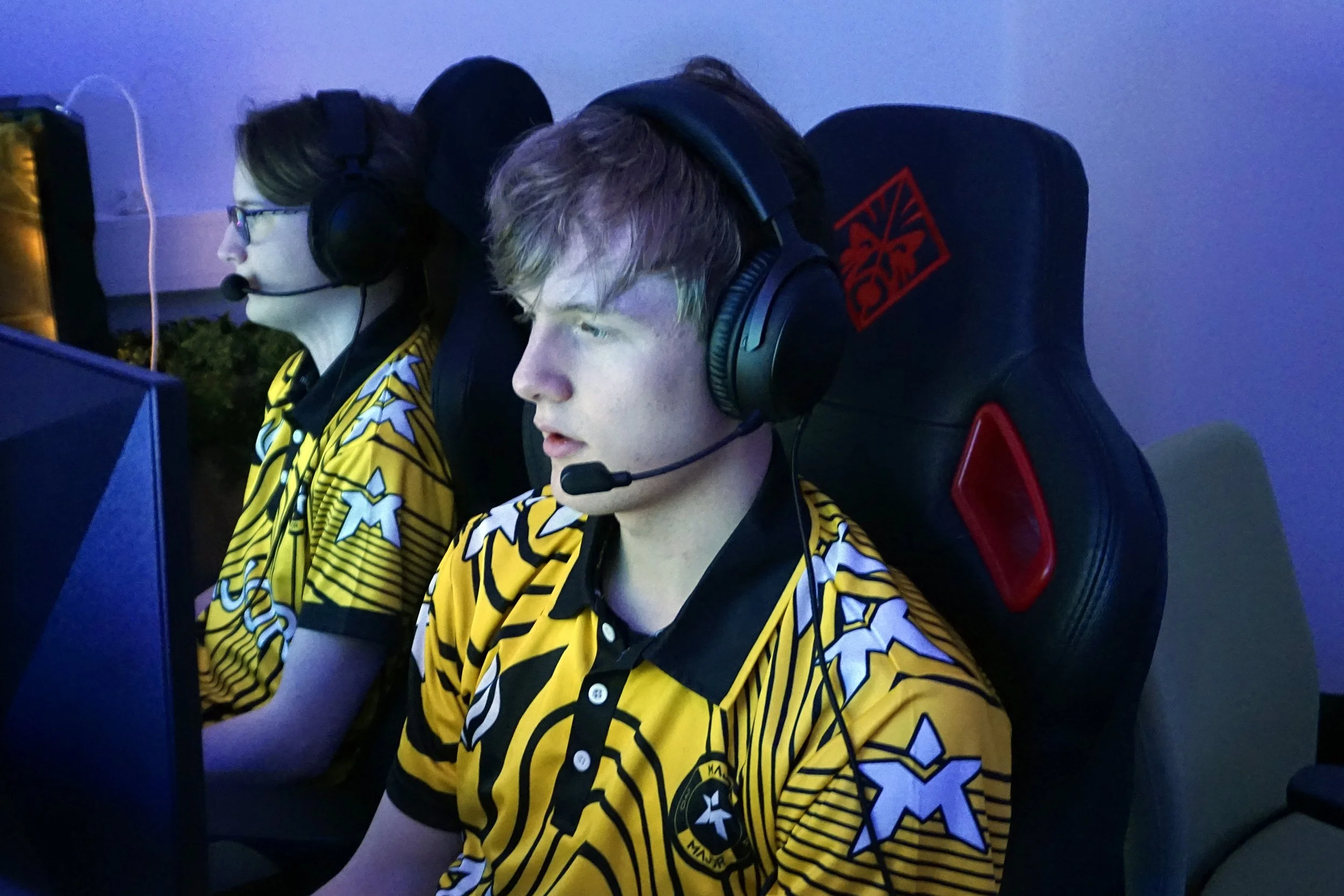 To personer i e-sportutstyr som spiller på datamaskiner, iført gul og svart lagdrakter med logoer, i et gamingrom.