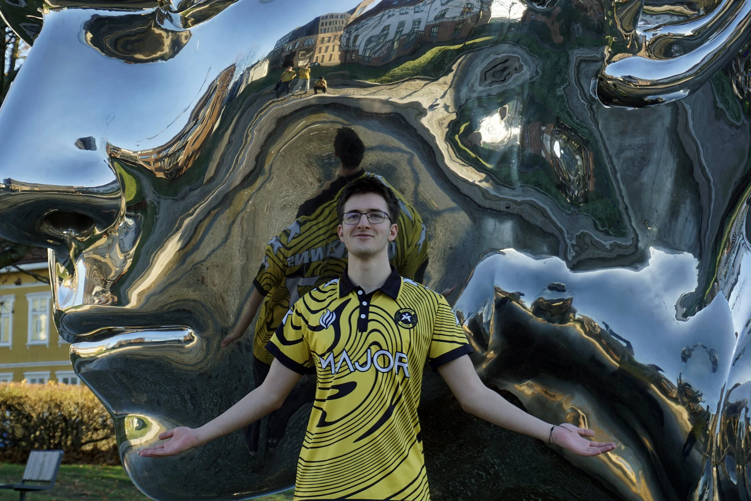 En ungdom foran en stor, skinnende, sølvfarget skulptur som reflekterer rundt om ham, med flere personer og bygninger reflektert i overflaten.