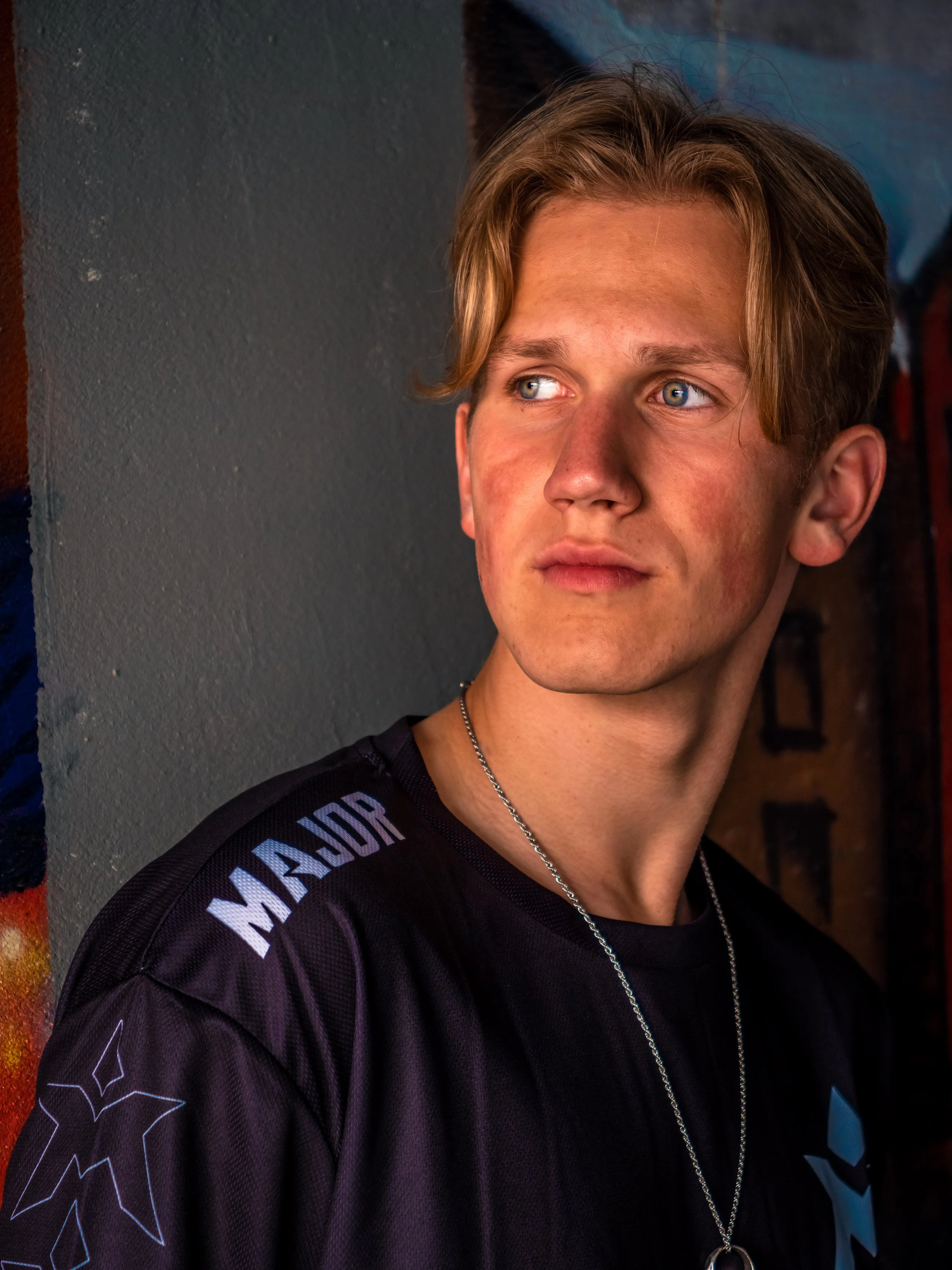 En ung mann med blond hår, iført svart sports-T-skjorte med logoer, står ved en vegg med graffiti. Han ser til siden med et tankefullt uttrykk.