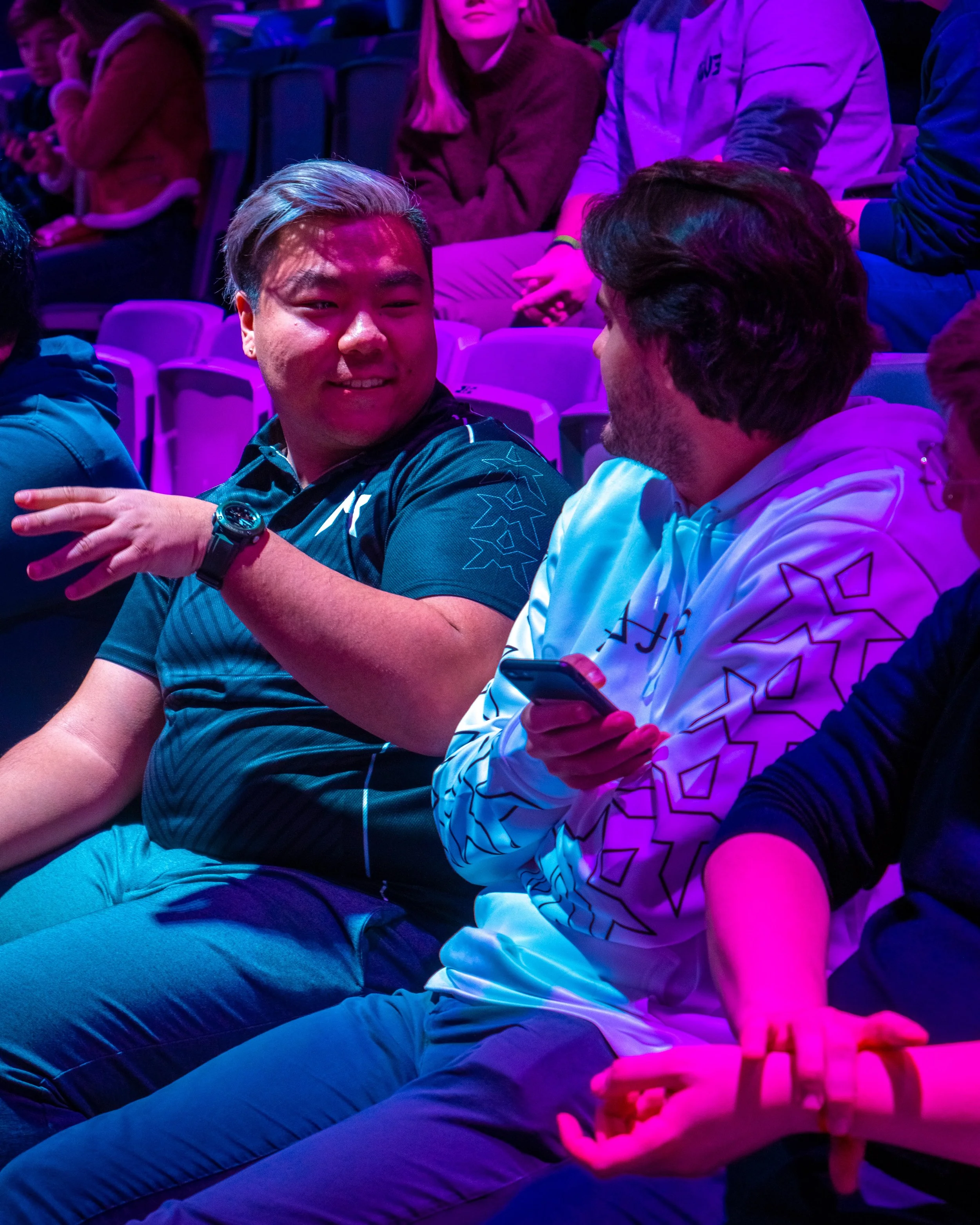 To menn som sitter og snakker sammen i et lyst arrangementsrom med andre mennesker rundt seg, hvorav noen ser på mobilen. Menneskene er omgitt av fargerik belysning.