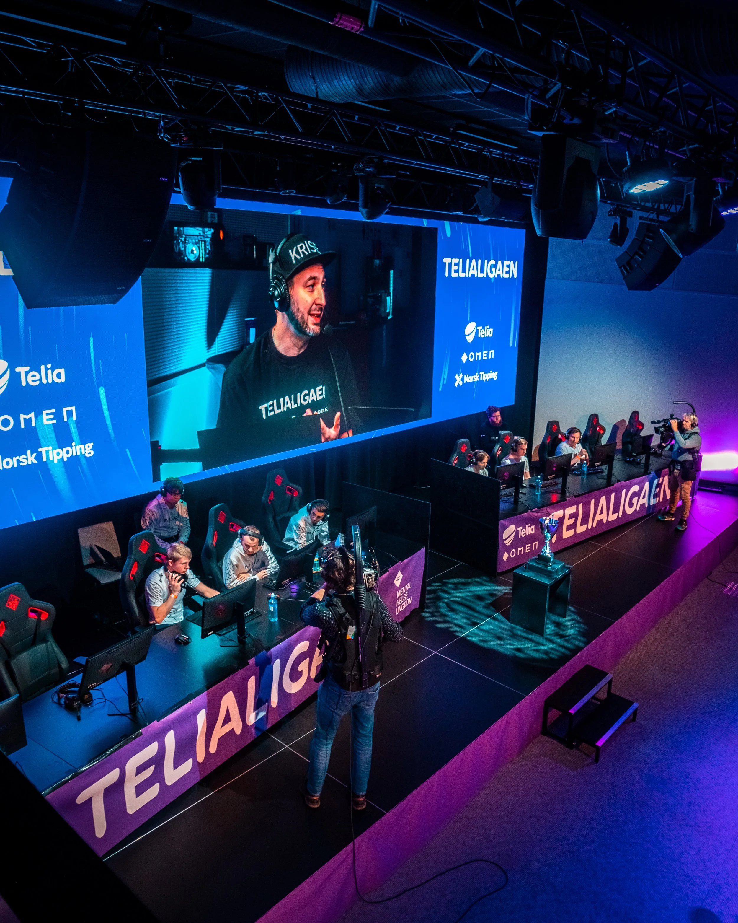 Et esportarrangement med lag som konkurrerer foran store skjermer. En person står og snakker til teamet, og en kameraoperatør filmer. Det er branding for Telia, Omen og Norsk Tipping.