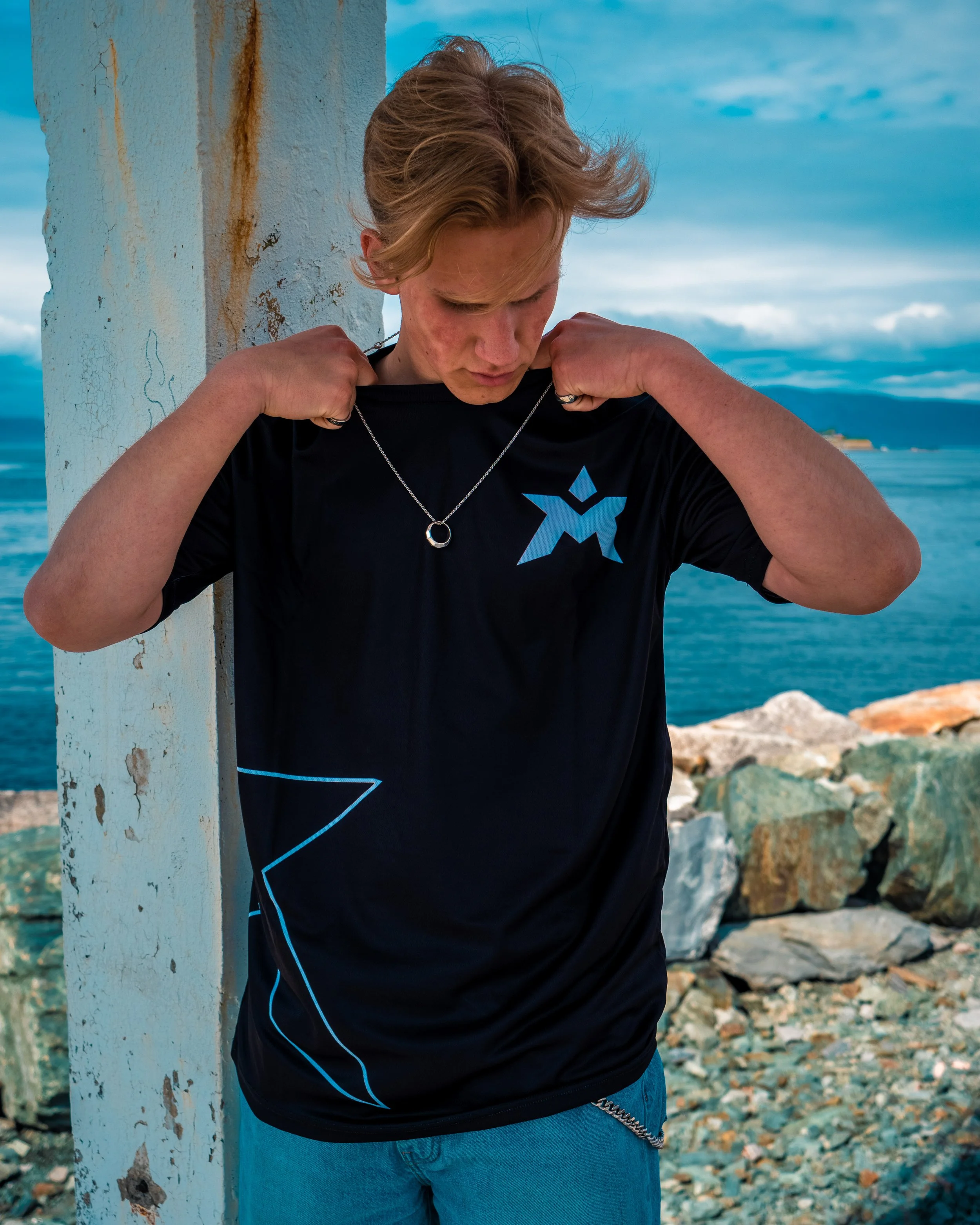 Ung mann med blond hår står ved en betongvegg ved sjøen, iført svart t-skjorte og jeans, og ser ned mens han justerer halskjede.