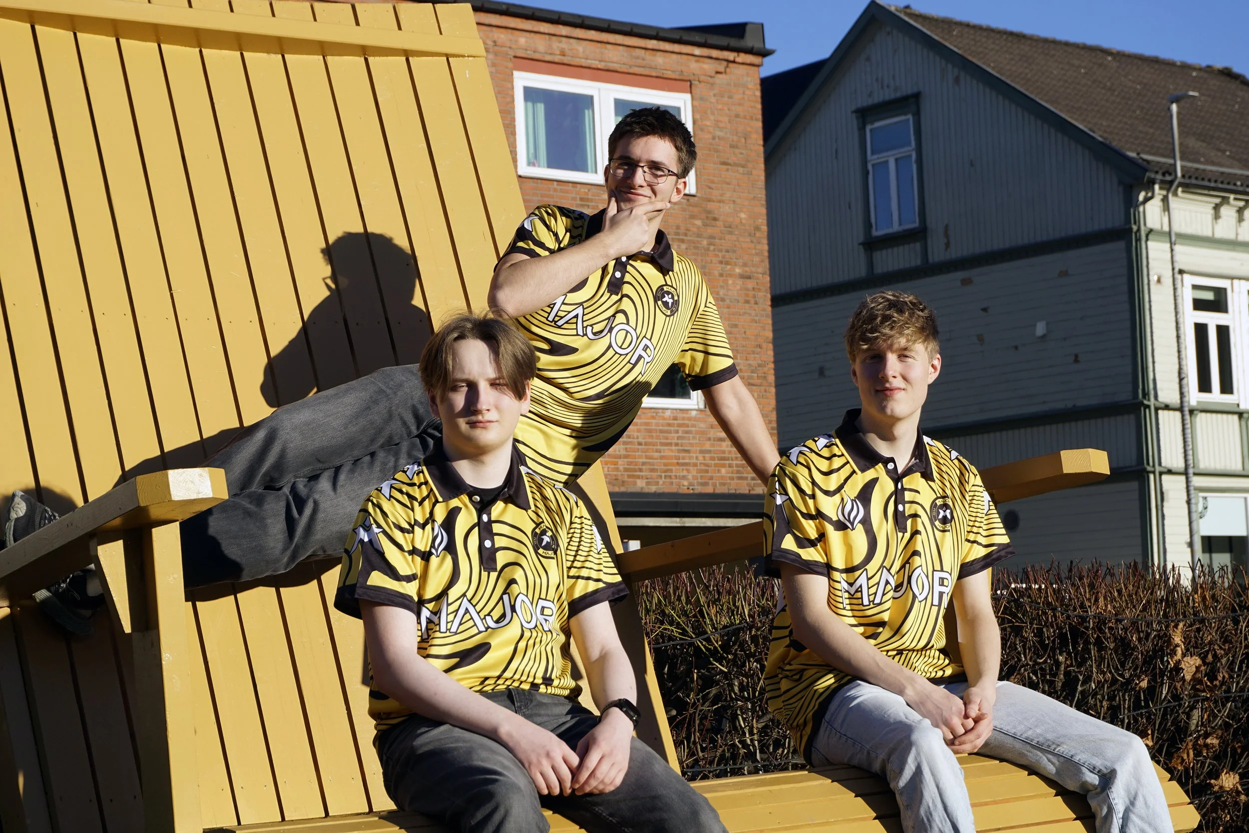 Tre unge personer i matchende gule og svarte sportsklær sitter på en stor, trebenk utendørs i solen, med en bakgrunn av bolighus og busker.