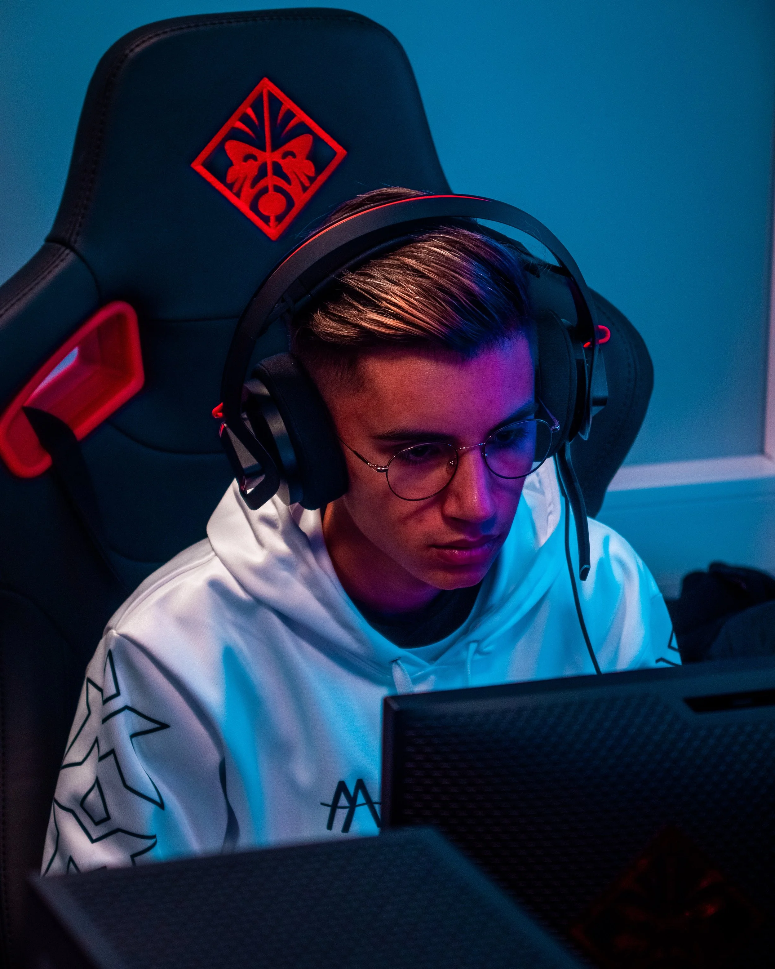 En ung mann med briller som spiller videospill foran en dataskjerm, sitter i en gamingstol med et kjempeskog-motiv på ryggen.