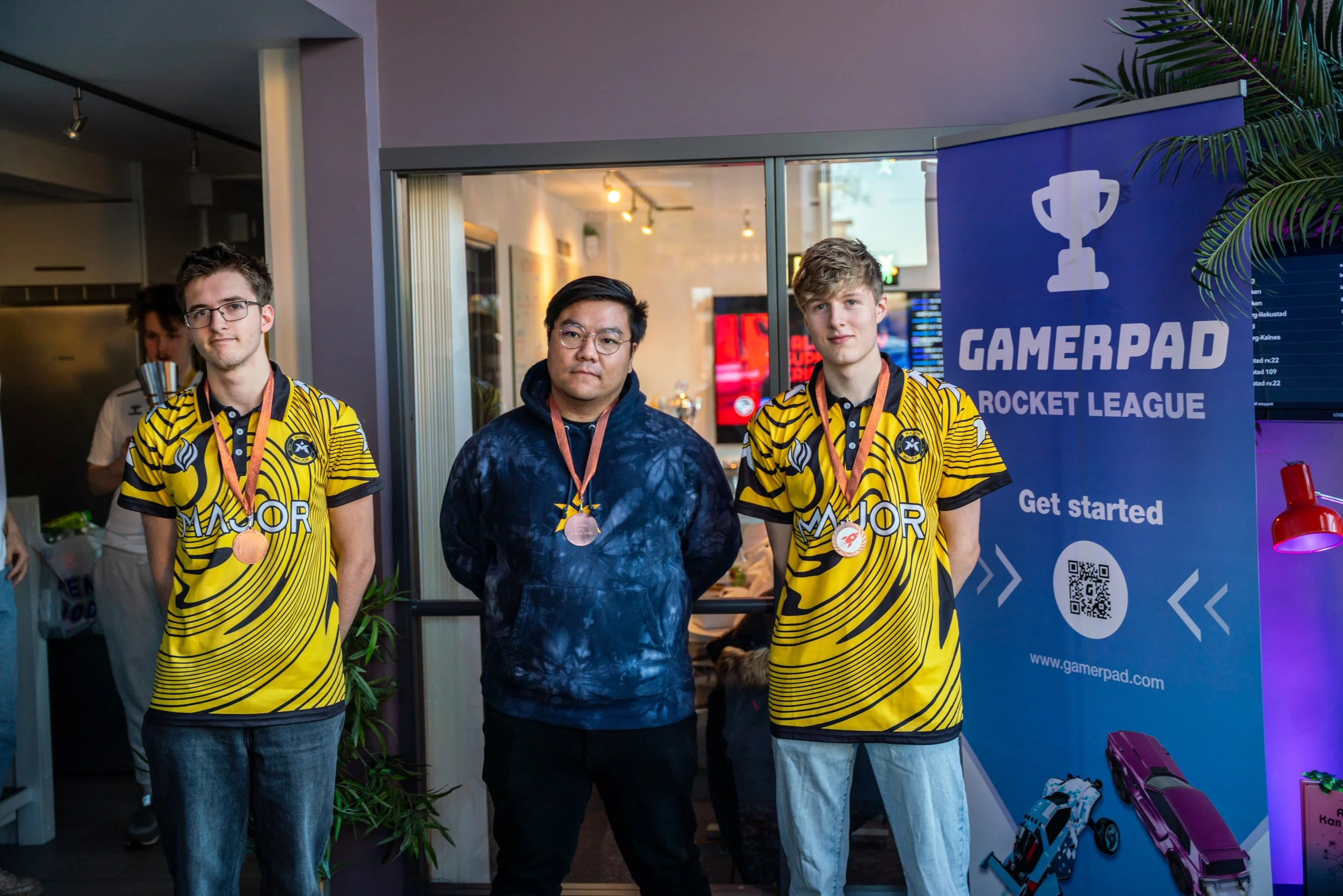 Tre ungdommer med brunt og blondt hår, to iført gult og svart jerseykleder med logoer og medaljer, står foran en Gamerpad Rocket League banner på en kafé eller arrangement, sammen med en annen person i blå jakke.