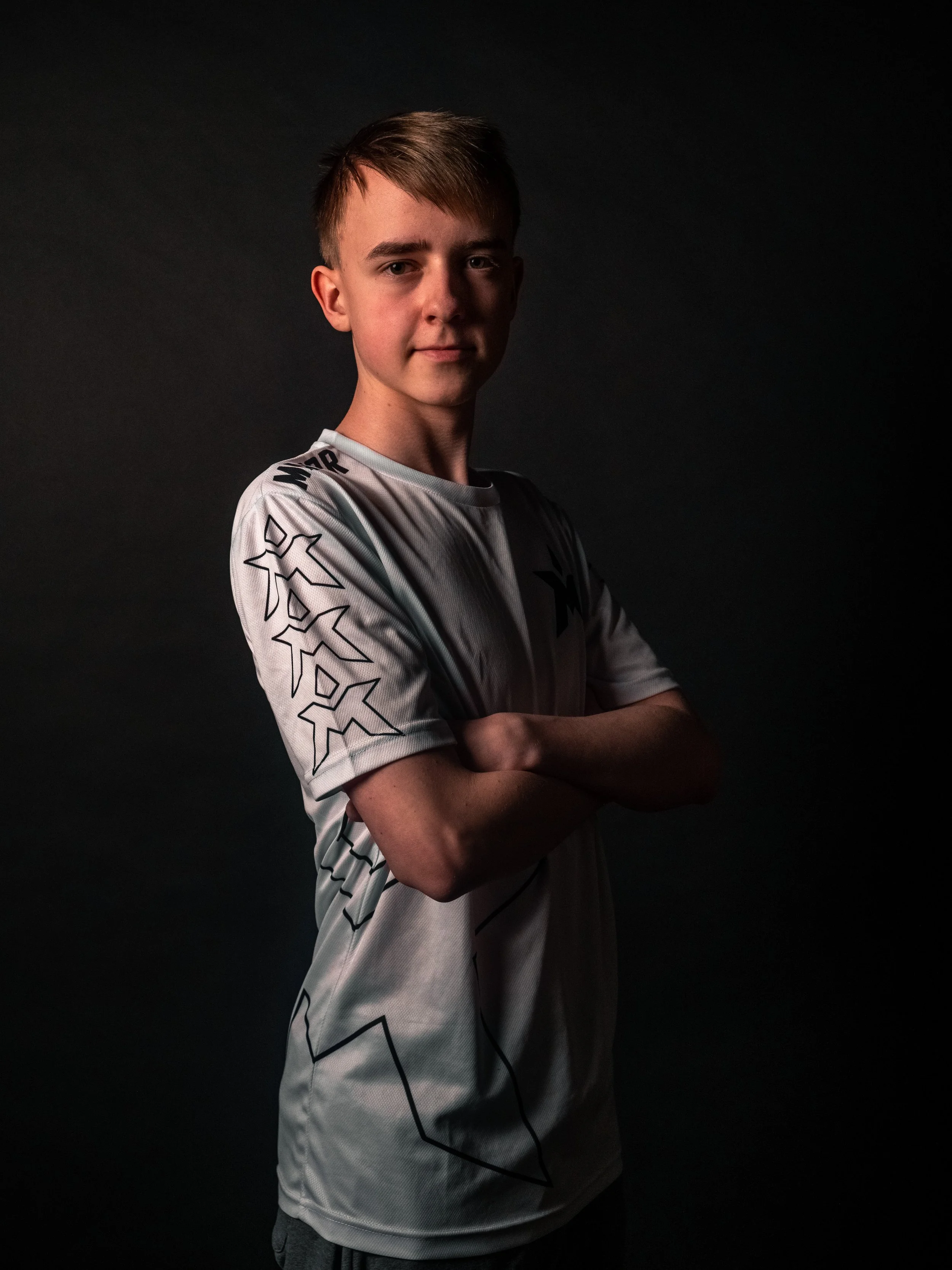 Portrett av en ung mann med kort hår, kledd i en hvit sports jersey med sorte grafiske mønstre, stående med armene korslagt mot en mørk bakgrunn.