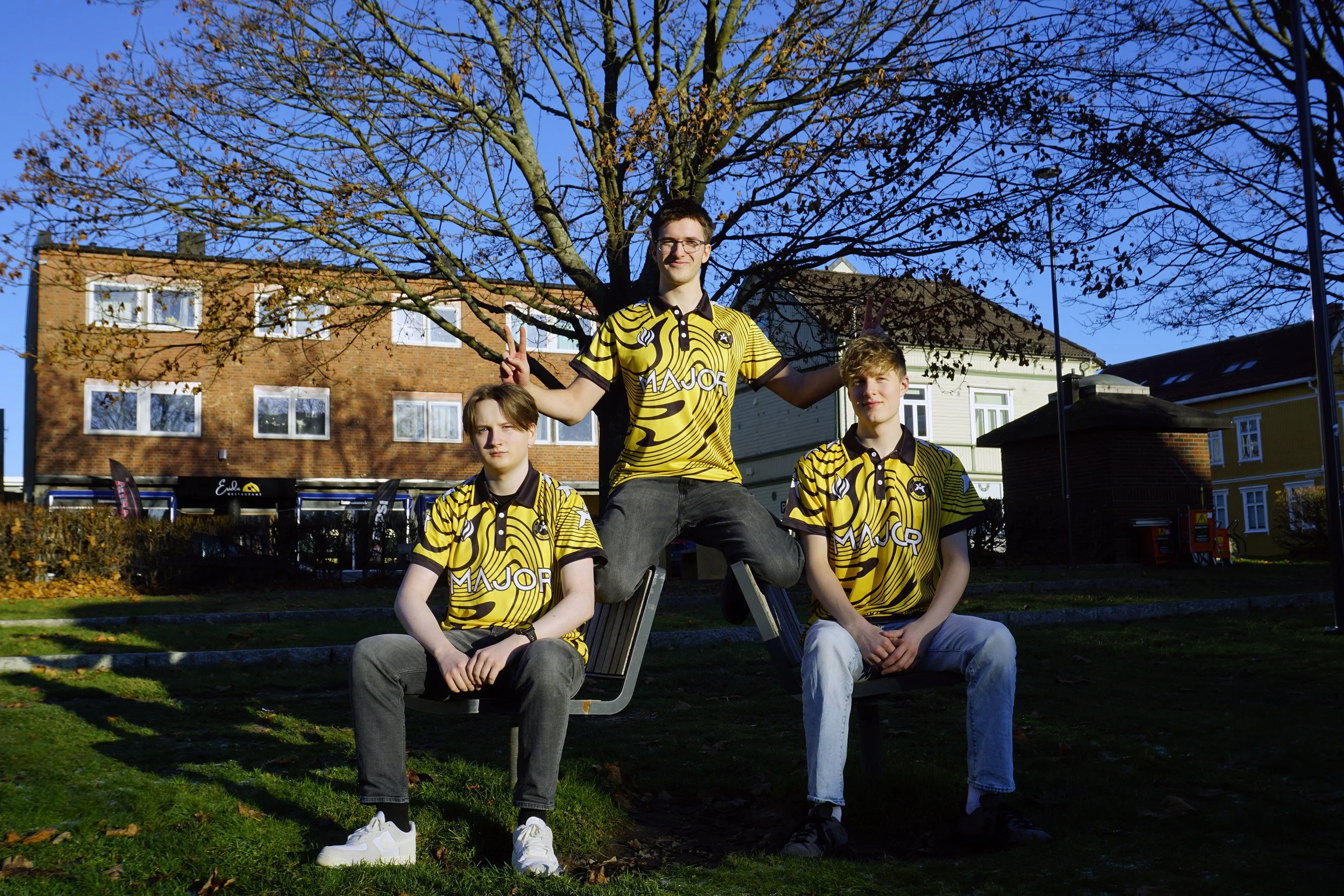 Tre ungdommer i gule og svarte sportsklær som sitter og står på en benk i en park med høstløv, trær og bygninger i bakgrunnen, og klar blå himmel.