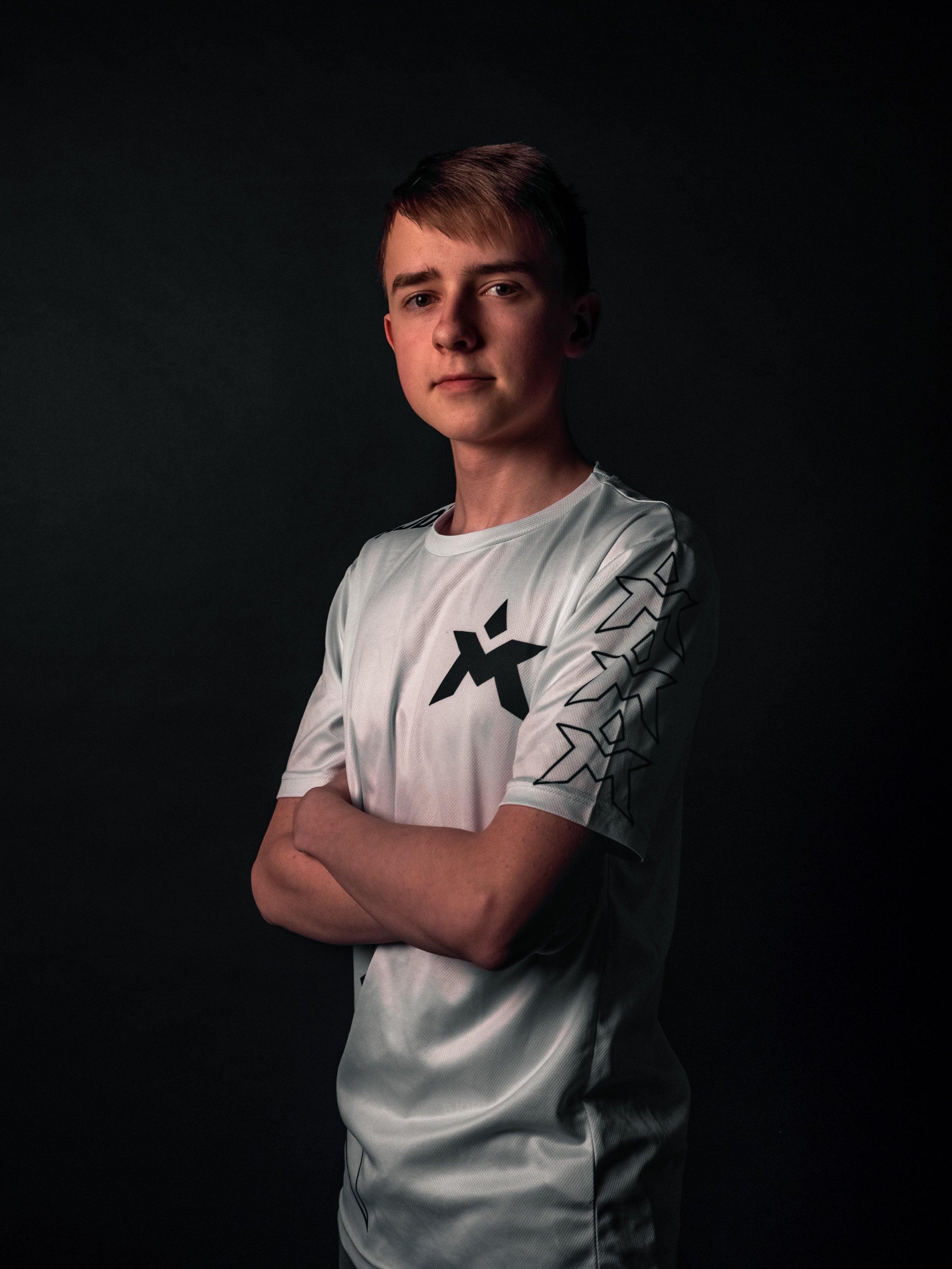 En ung mann med kort, brunt hår som står opp, kledd i en hvit sports t-skjorte med svarte grafiske mønstre på ermene, står med armene krysset mot en mørk bakgrunn.