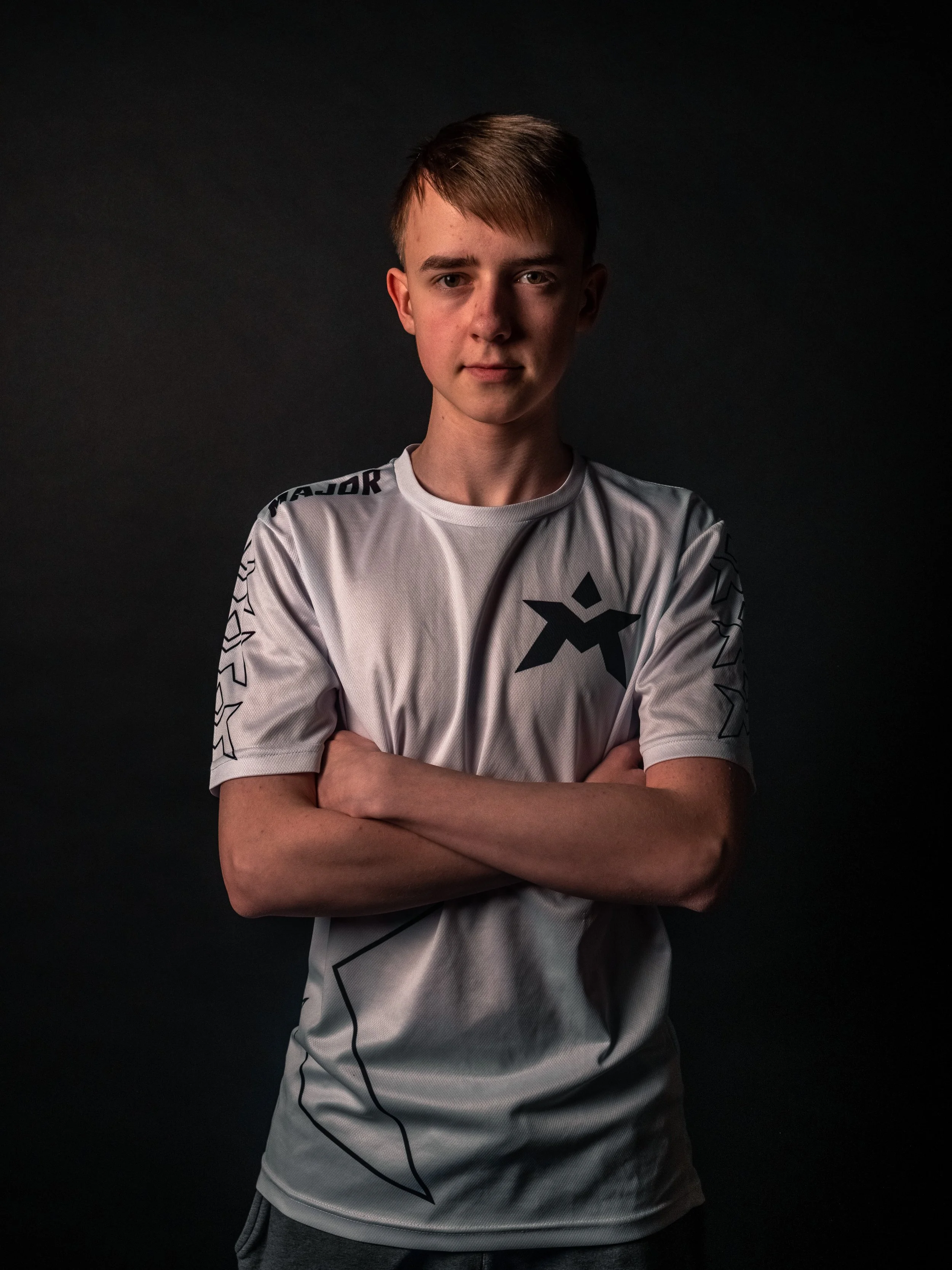 En ung mann står med armene overkors, iført en hvit sportskjorte med svarte logoer og designs, mot en mørk bakgrunn.
