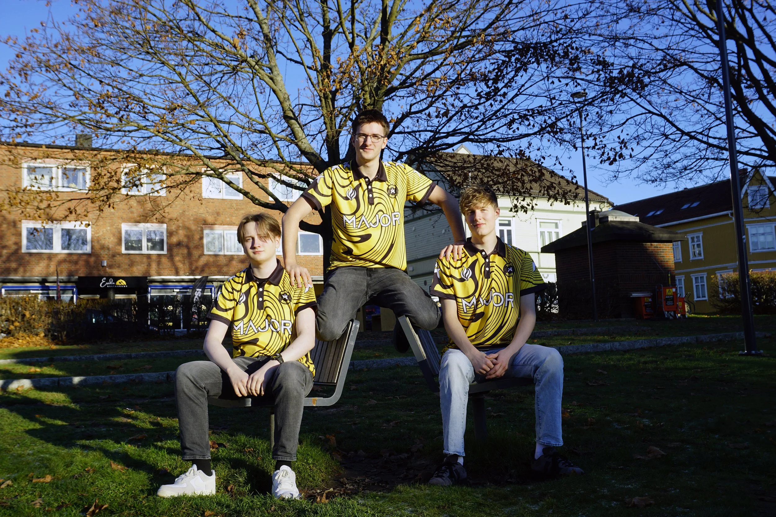 Tre unge menn i gule og svarte sportsjerseyer og jeans, to sitter på benker, den tredje står med foten på benken, foran hus og trær med høstblad, under en klar blå himmel.
