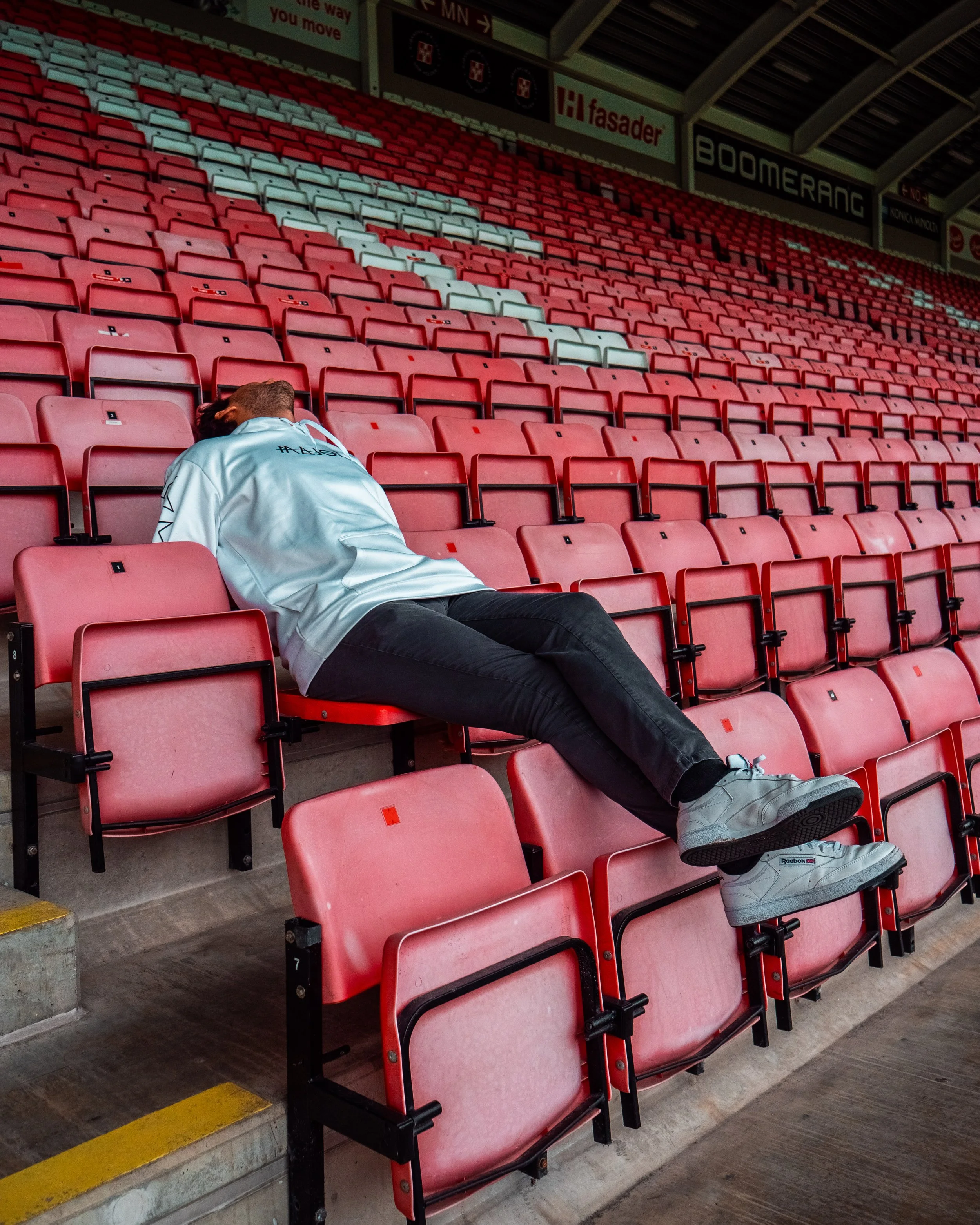 En person ligger utstrakt på en tom tilskuerplass i et fotballstadion, omgitt av røde seter.