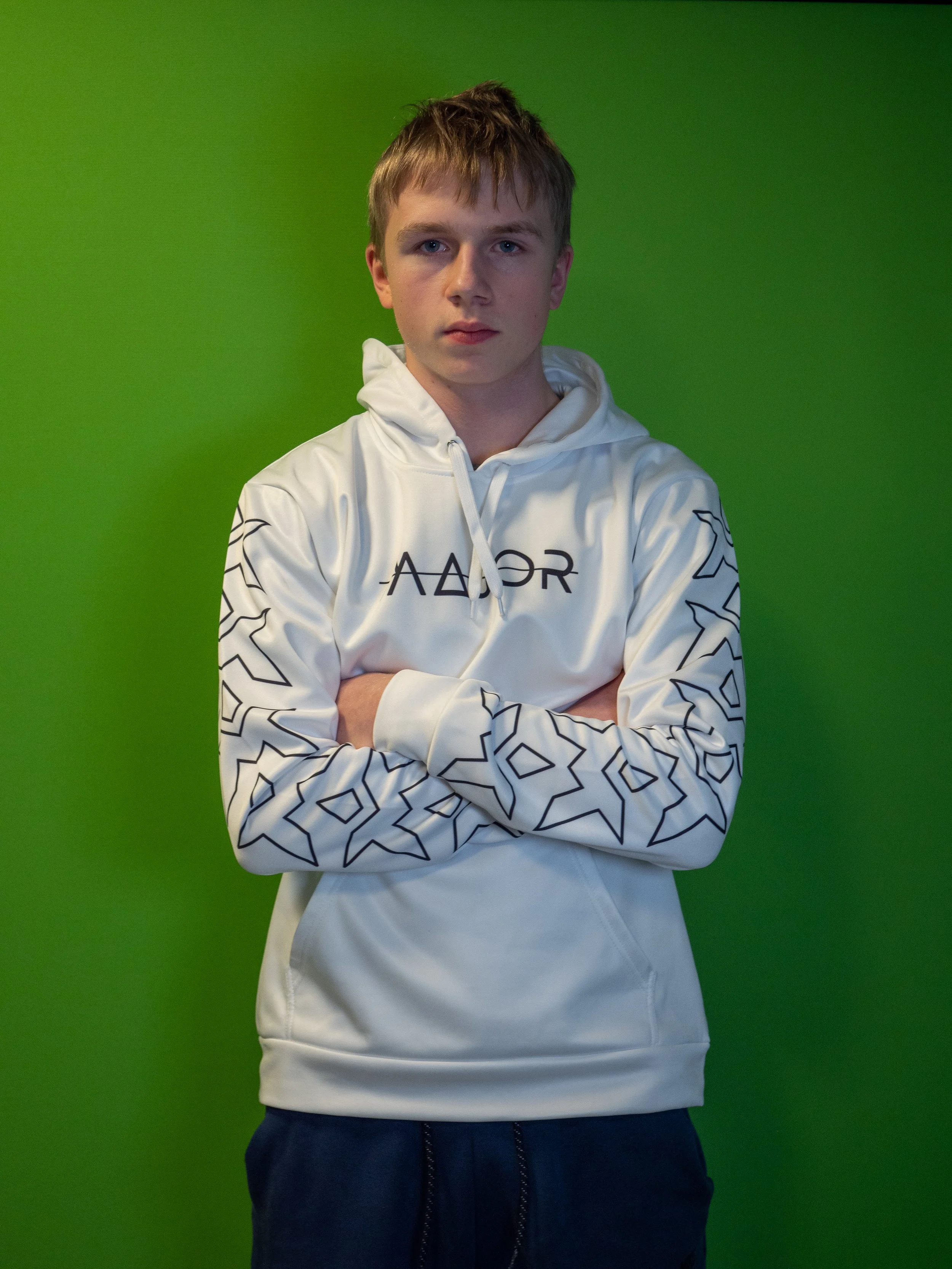 Ung mann står med armene i kors foran en grønn bakgrunn, iført en hvit hettegenser med mønster på ermet.