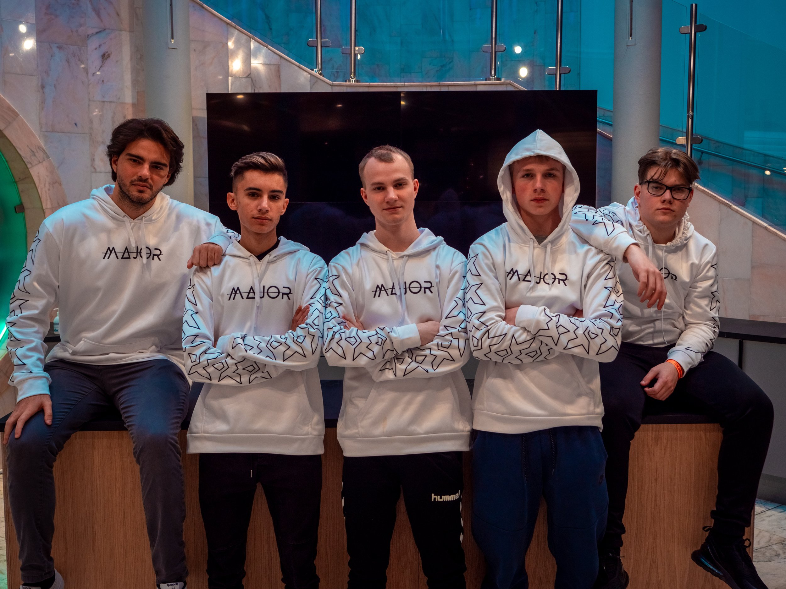 Fem unge menn som står på en benk innendørs, alle iført matchende hvite hettegensere med ordet 'Major' på.