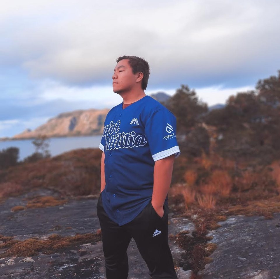 En ungdom iført en blå baseball-trøye står på en steinete mark med hender i lommene, ser ut til å nyte naturen med bakgrunn av fjell og vann under en overskyet himmel.