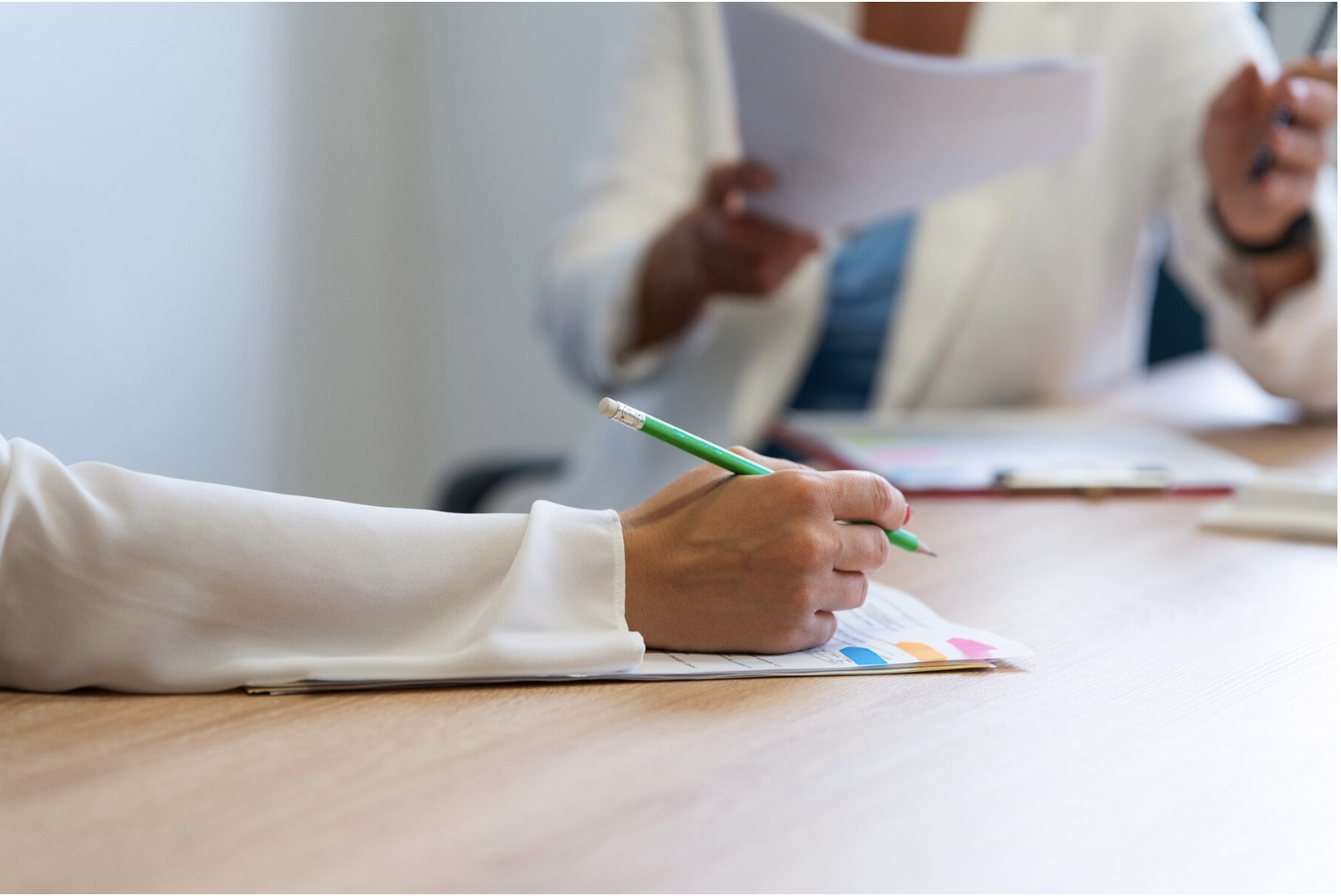 Une main tenant un stylo vert écrit sur un document à une table, avec deux autres personnes en arrière-plan tenant des papiers.