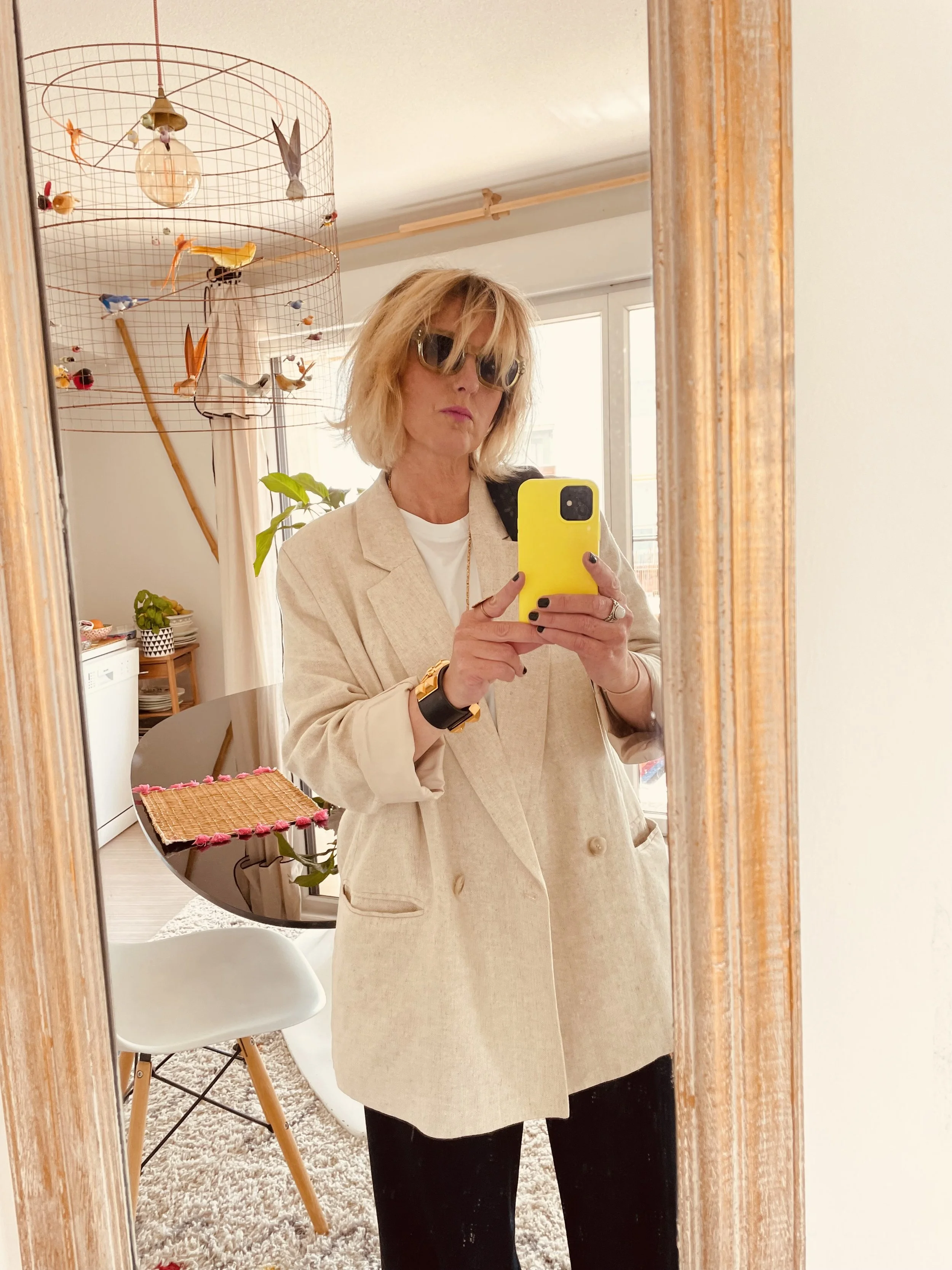 Femme prenant un selfie dans un miroir avec un téléphone jaune. Elle porte une veste beige, des lunettes de soleil et une montre noire et or. La pièce a une décoration moderne avec une lampe de plafond artistique, un arbre vert à l'arrière, une table en bois avec un napperon rose, et une chaise blanche avec des pieds en bois.