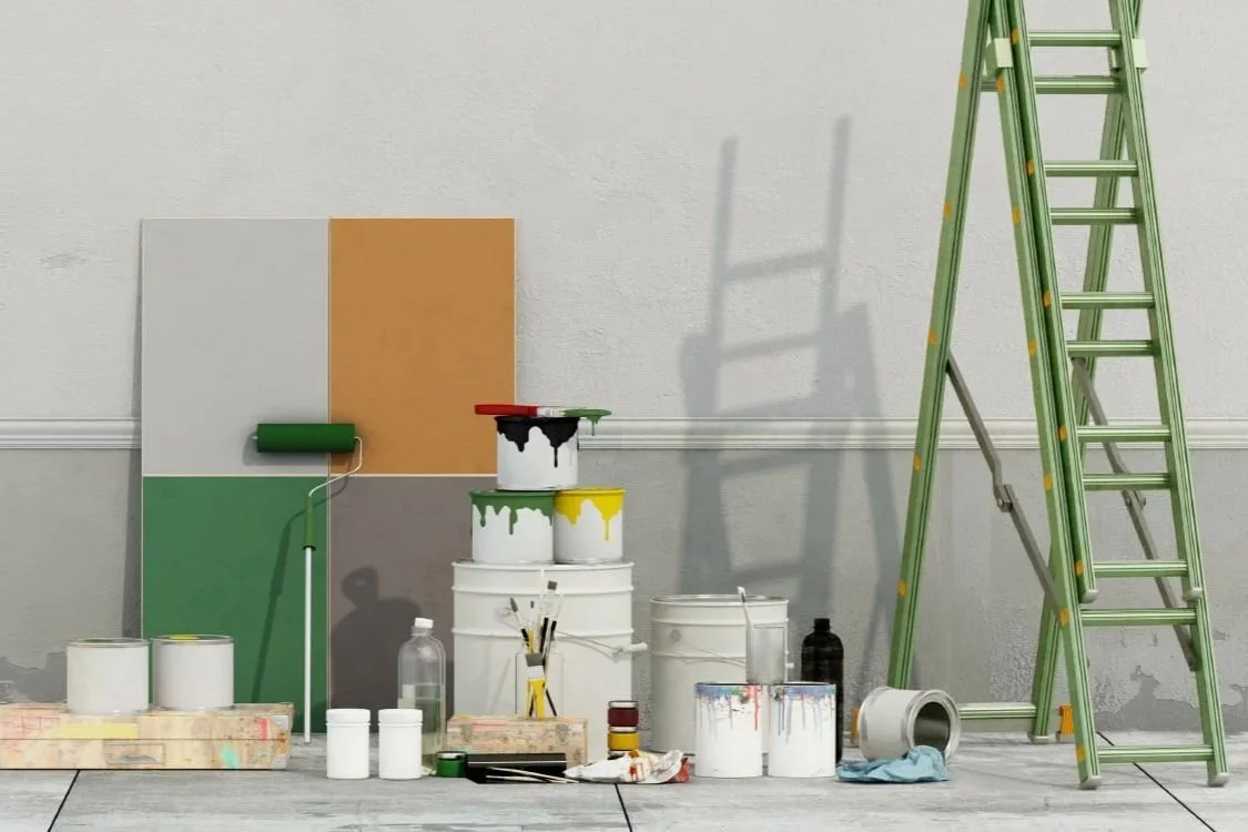 Painting supplies in a room with a ladder, paint cans, paint rollers, brushes, and painted canvas panels